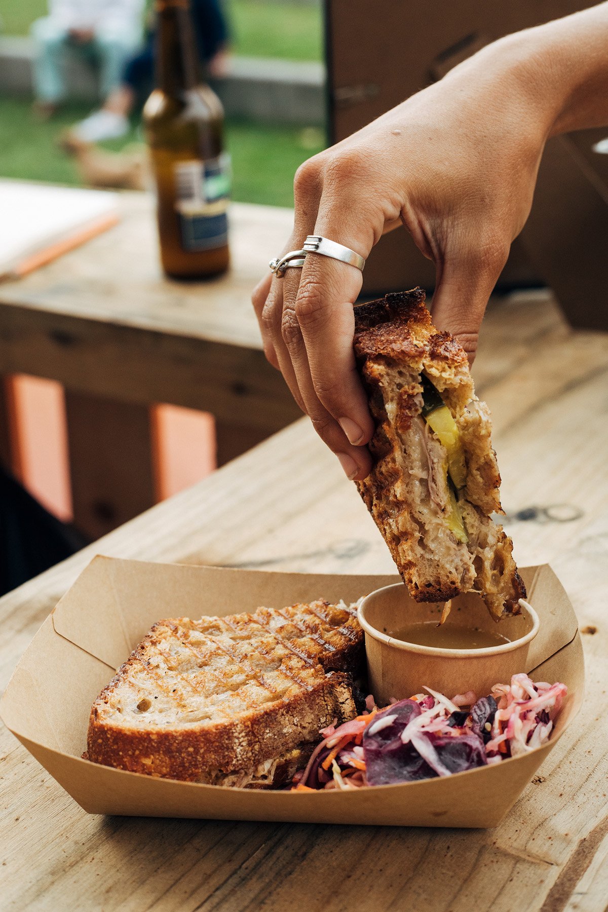 Main tenant un sandwich garni dans un panier en carton sur une table en bois — photographie culinaire pour marque street food ou contenu food branding.