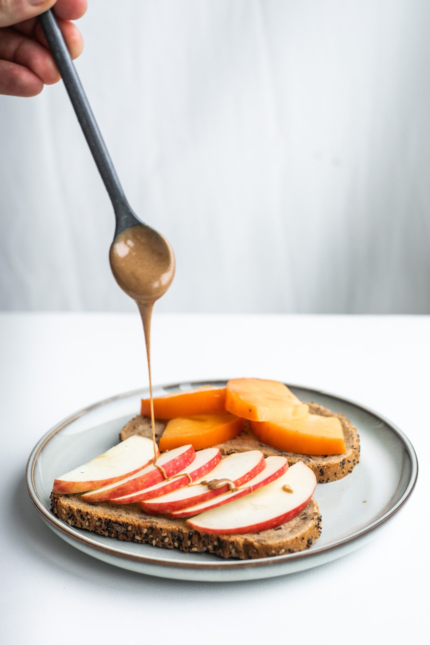 Photographie culinaire d’une main versant du sirop sur des tranches de pain aux céréales garnies de pommes et de poires.