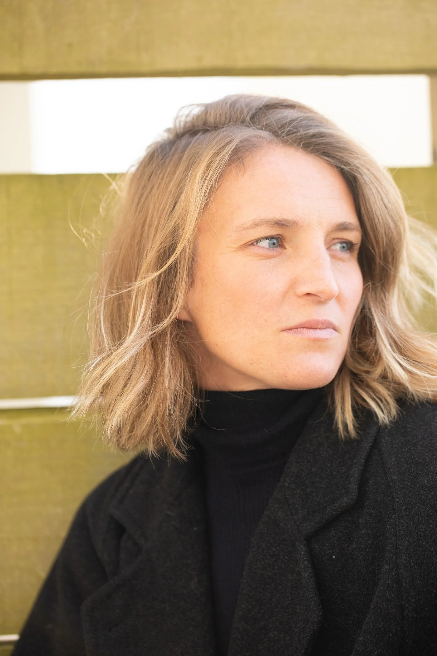 Portrait d’une femme blonde vêtue de noir, debout devant un mur vert olive — image branding personnelle en lumière naturelle.