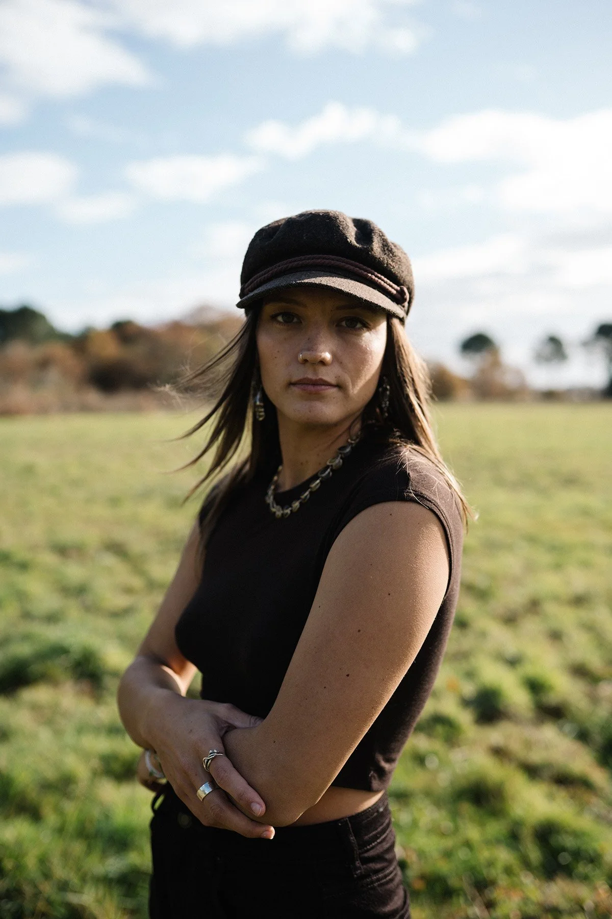 Portrait d’une jeune femme en tenue noire et casquette, debout dans un champ sous ciel nuageux — image de marque personnelle avec ambiance contemplative.