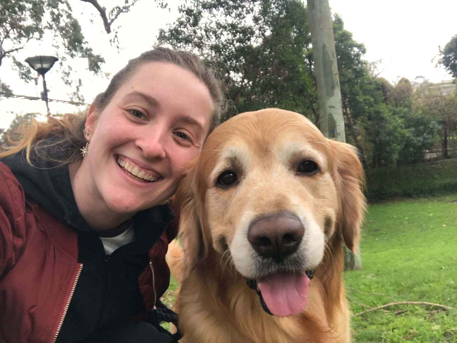 A woman smiling with a happy expression taking a selfie with a golden retriever dog in an outdoor park setting with trees and grass.