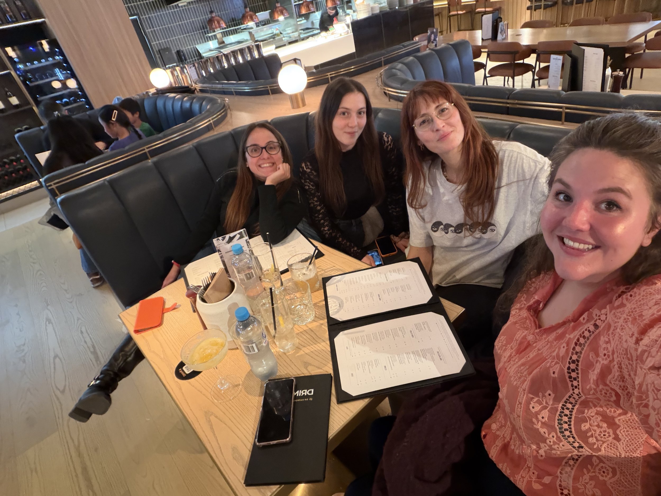 Four women sitting at a restaurant table, smiling and taking a selfie. On the table, there are drinks, a menu, and condiments, with a busy restaurant in the background.