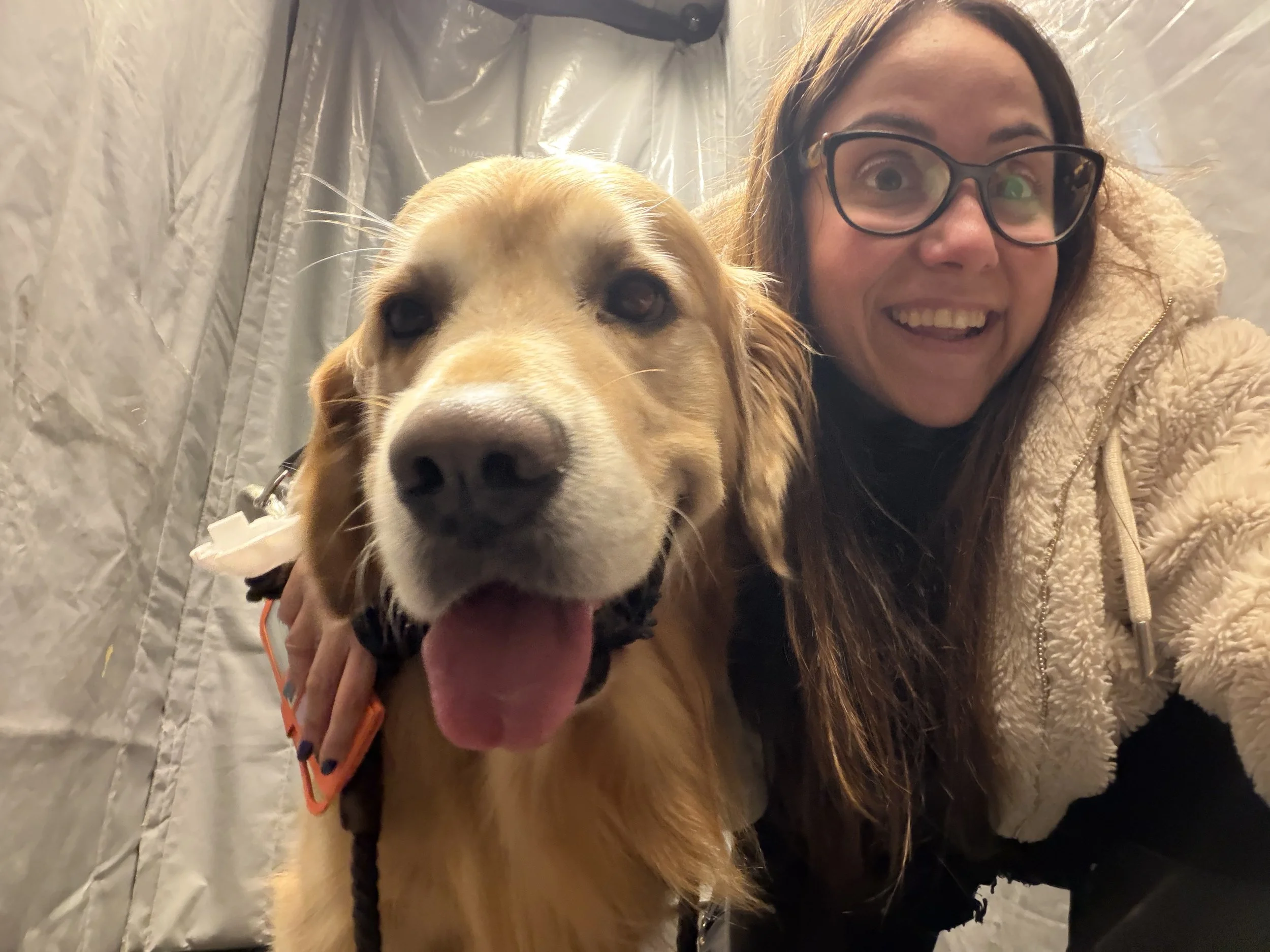 A woman with long brown hair, glasses, and a beige fluffy jacket smiling with her pet dog, a large golden retriever, inside a metallic or shiny enclosed space.