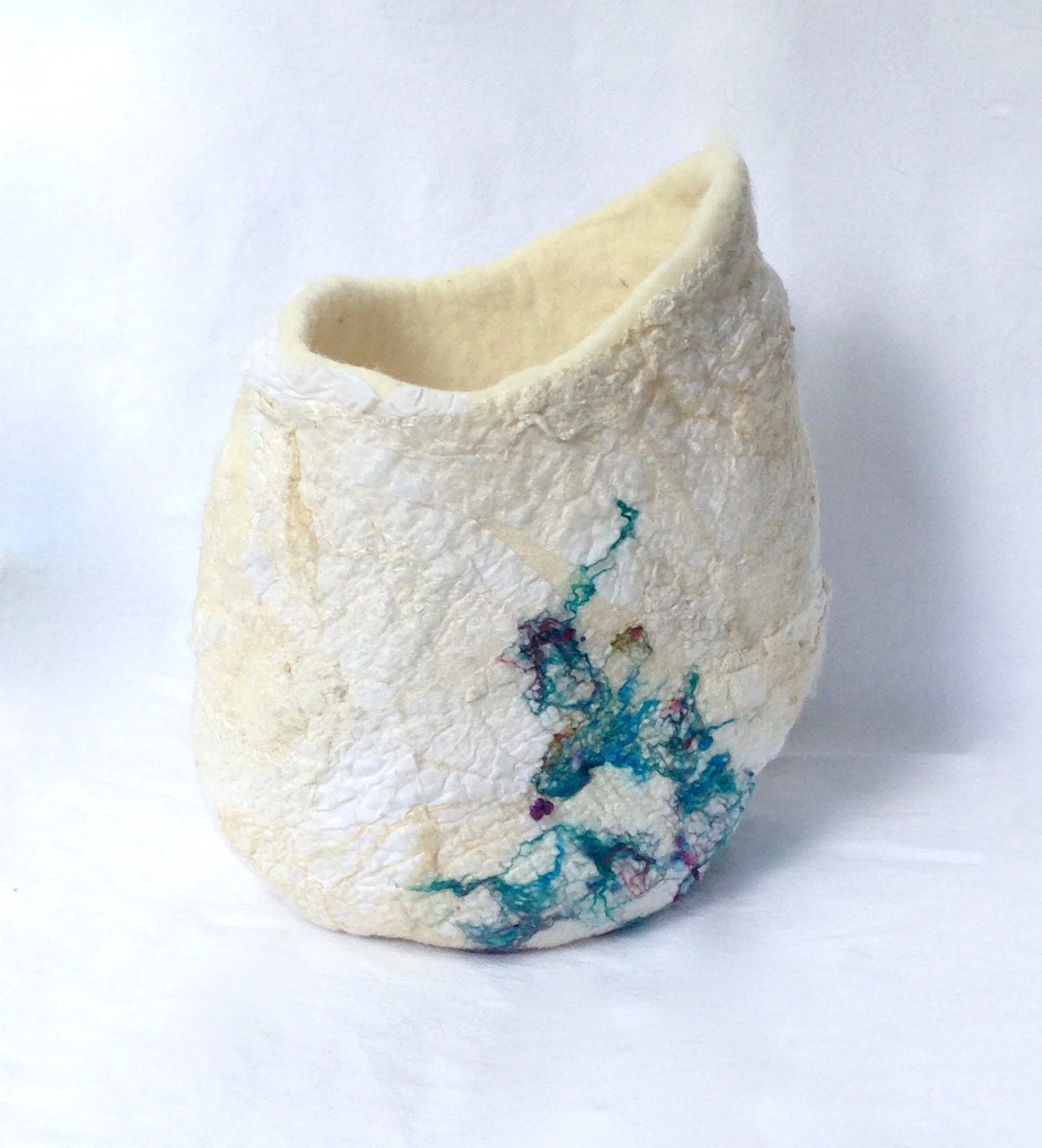 A felt vessel with an irregular shape, cream-colored surface, and colorful thread-like design at the bottom, set against a plain white background.
