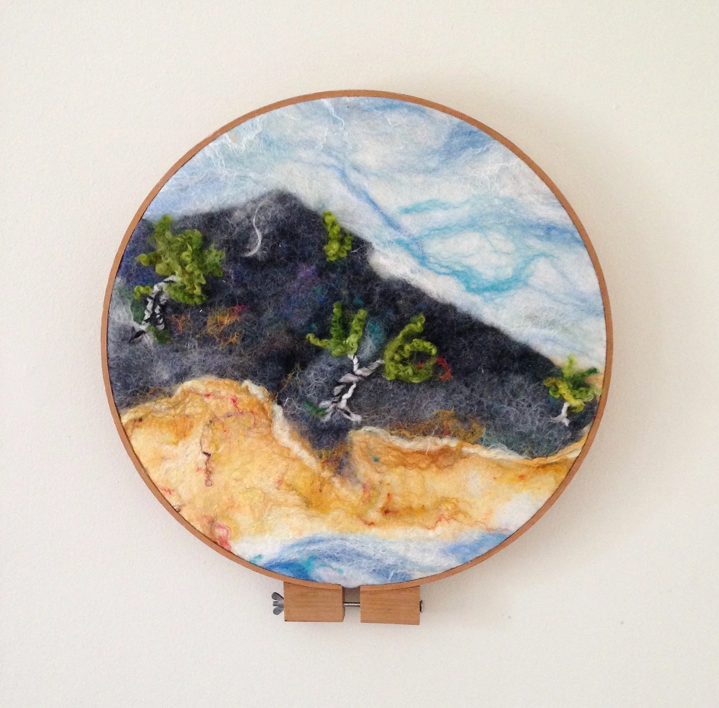 Felted landscape art depicting mountains, trees, a sky with clouds, and a sandy foreground within a circular frame.
