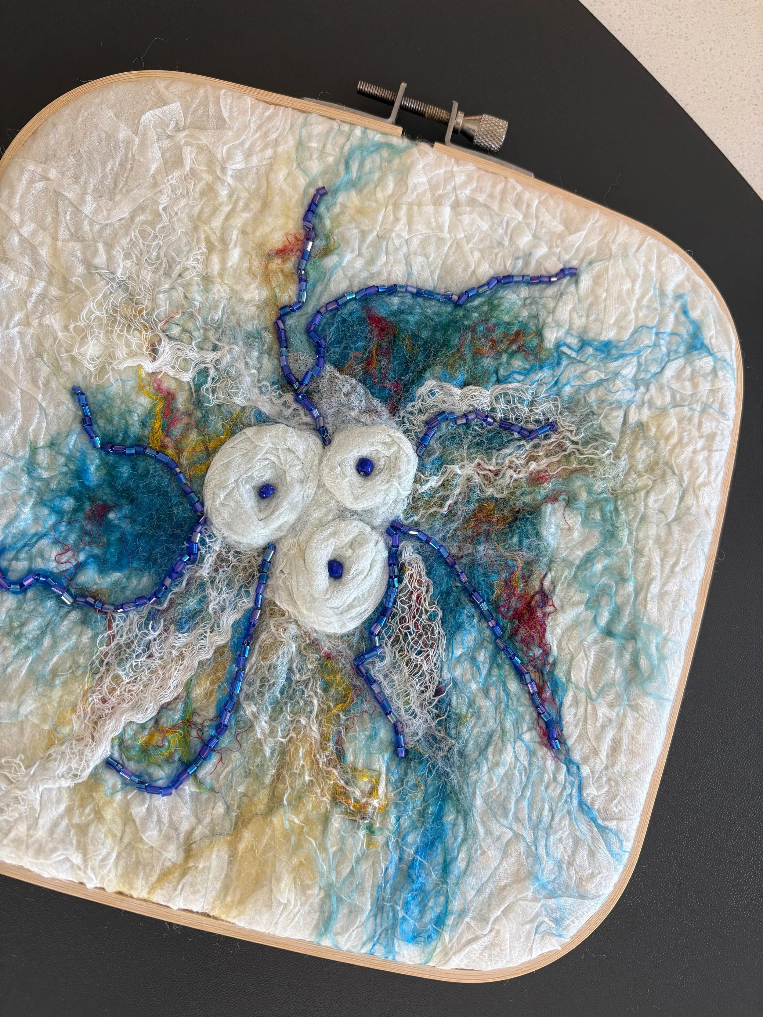 Close-up of the embroidery hoop-framed felt in shades of blue and white with intricate thread details, beads, and colorful accents.