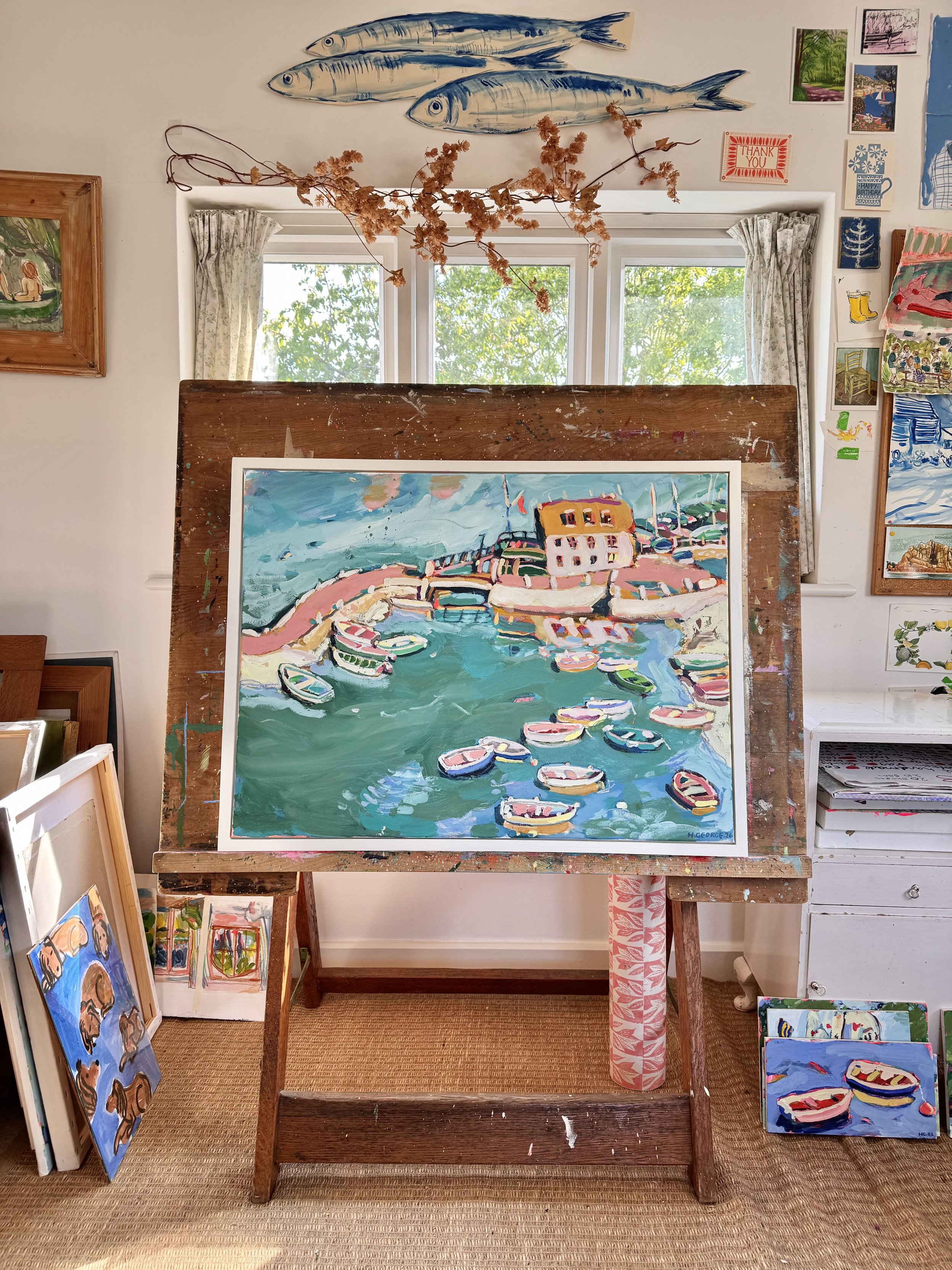 An art studio with paintings and drawings, featuring a prominent colorful painting of a harbor scene with boats and buildings, displayed on an easel. The studio has small fish sculptures on the wall, a branch with dried flowers hanging, and various a