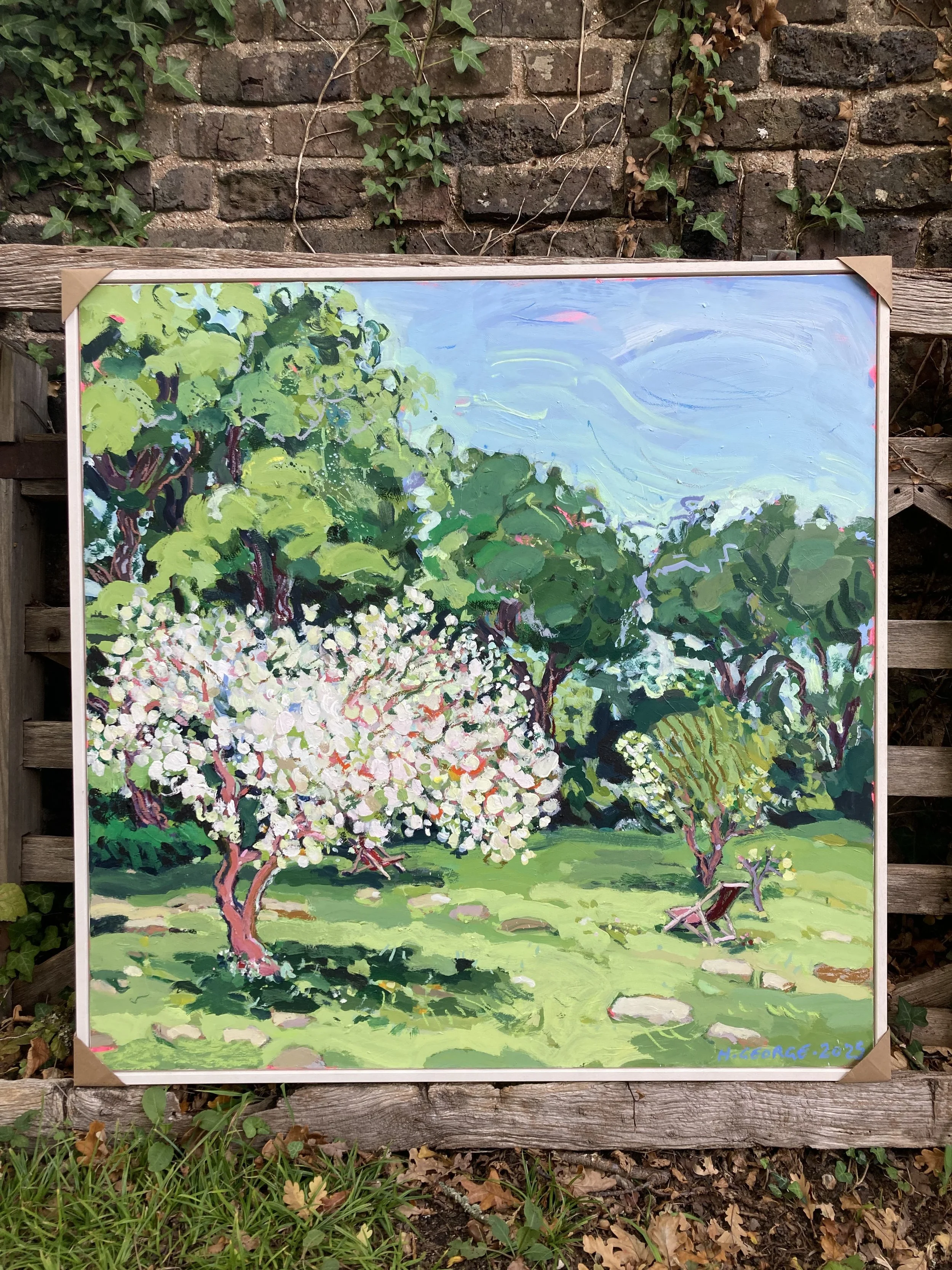 A colorful landscape painting showing trees with white blossoms, green foliage, and a blue sky, framed and leaning against a wooden fence with ivy growing on it.