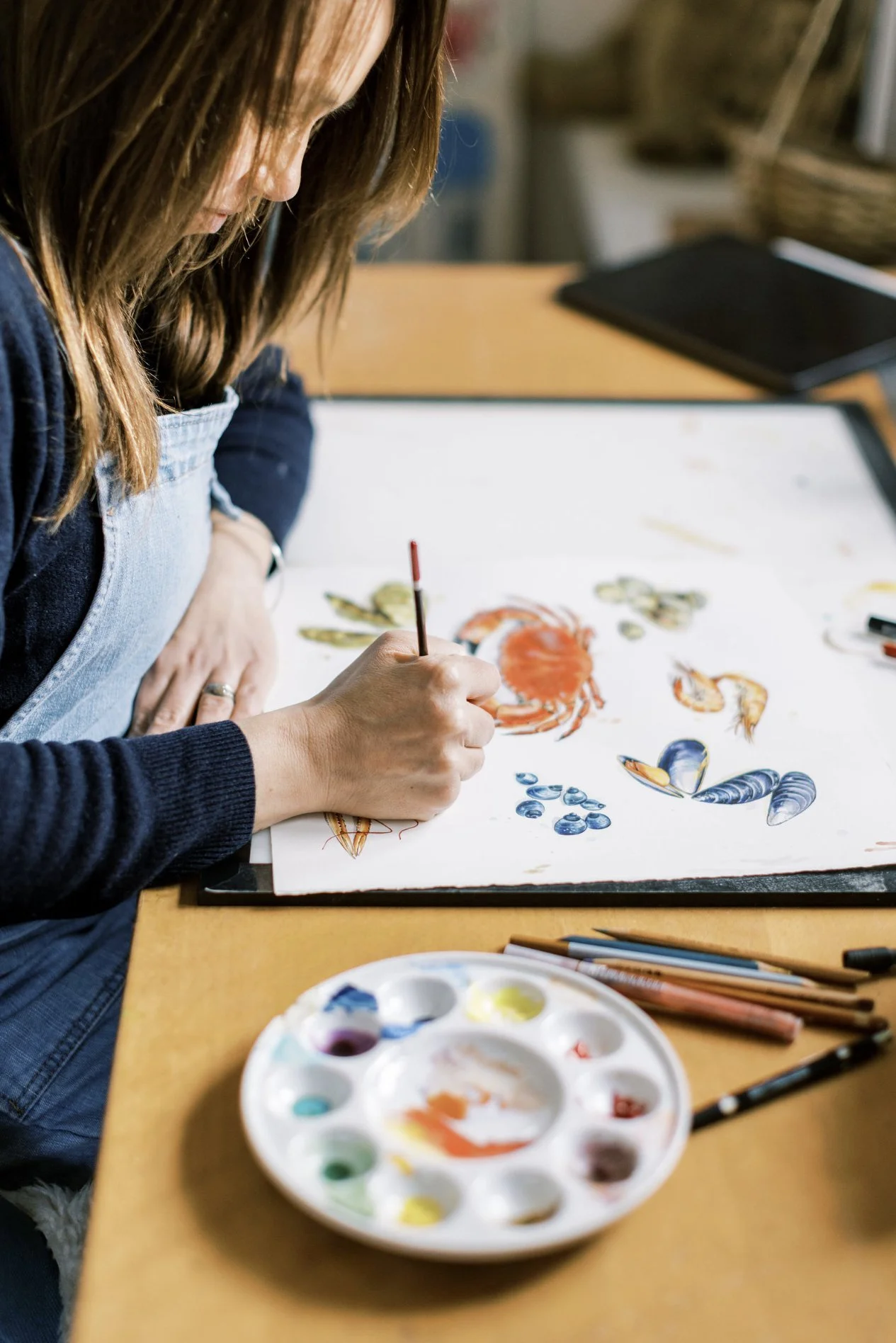 Hannah George painting a detailed watercolour of sea creatures, including a crab, lobsters, and shells, on a white sheet of paper. The table holds a palette with various watercolour paints and several paintbrushes.