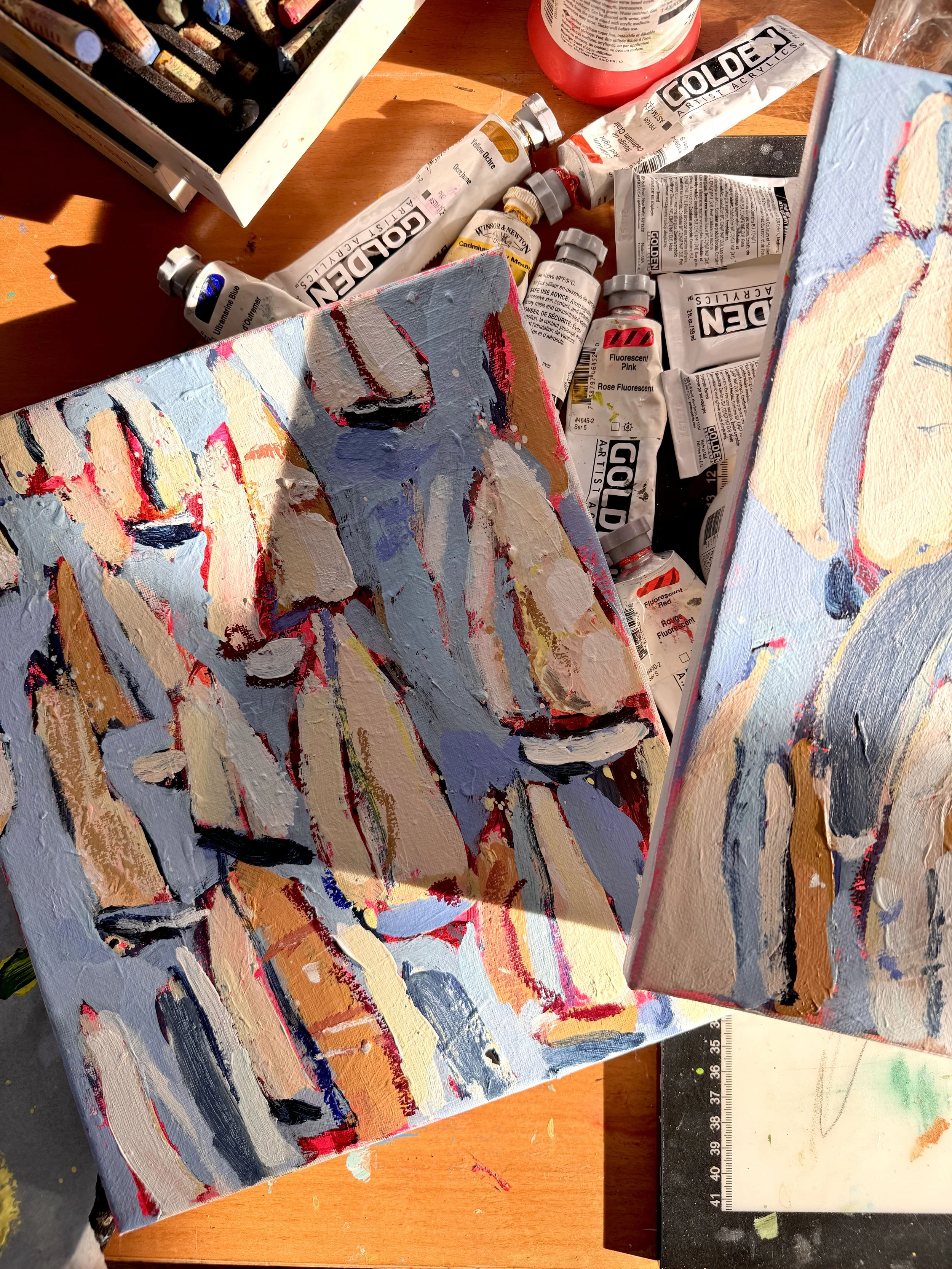 Paint tubes and a partially painted abstract canvas on a wooden table with a ruler in the background.