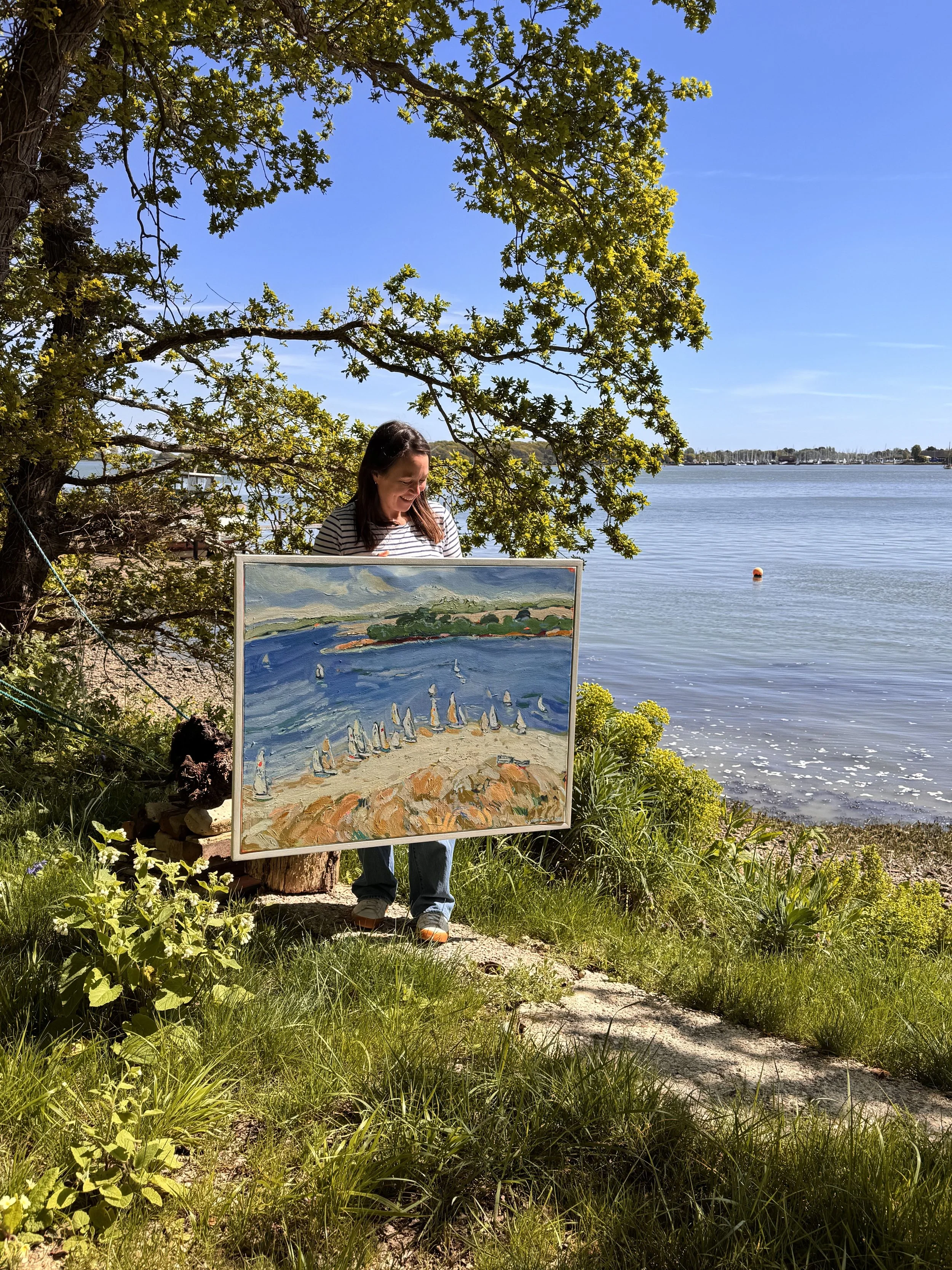 A woman stands outdoors near a lake, holding a painting of sailboats on the water. She smiles as she looks at the artwork, surrounded by green grass, bushes, and a tree with yellow flowers. The sky is blue with a few wispy clouds, and mooring buoys a