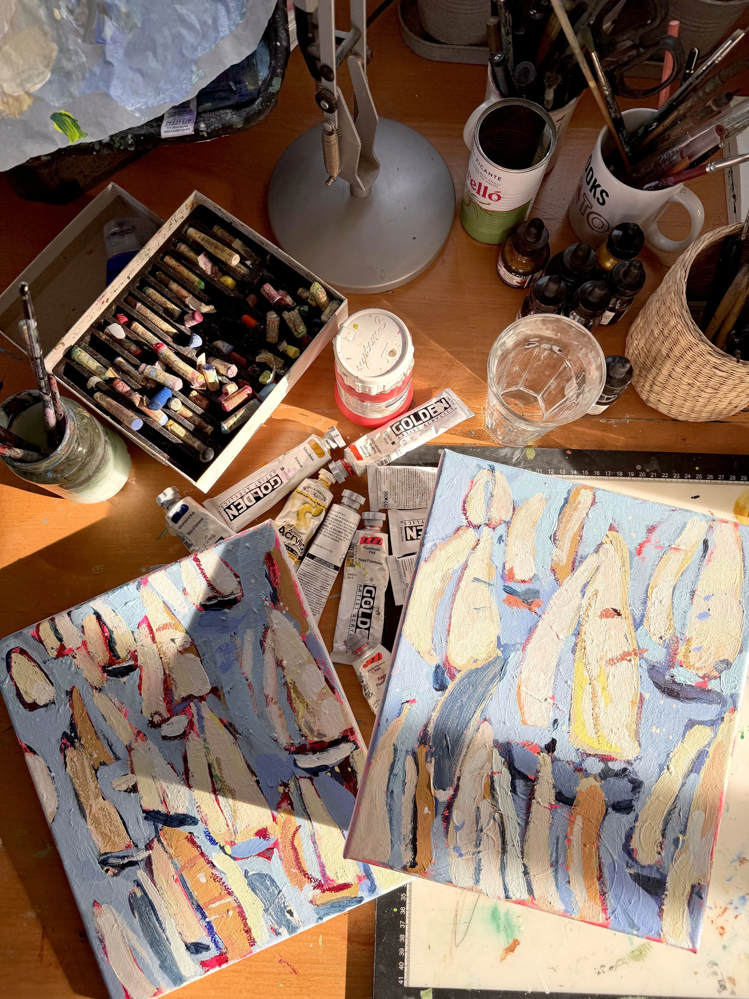 Two abstract paintings with colorful brushstrokes on paper, surrounded by art supplies including paint tubes, brushes in cups, a jar of water, and a palette with paint. The workspace is well-lit and artistic.