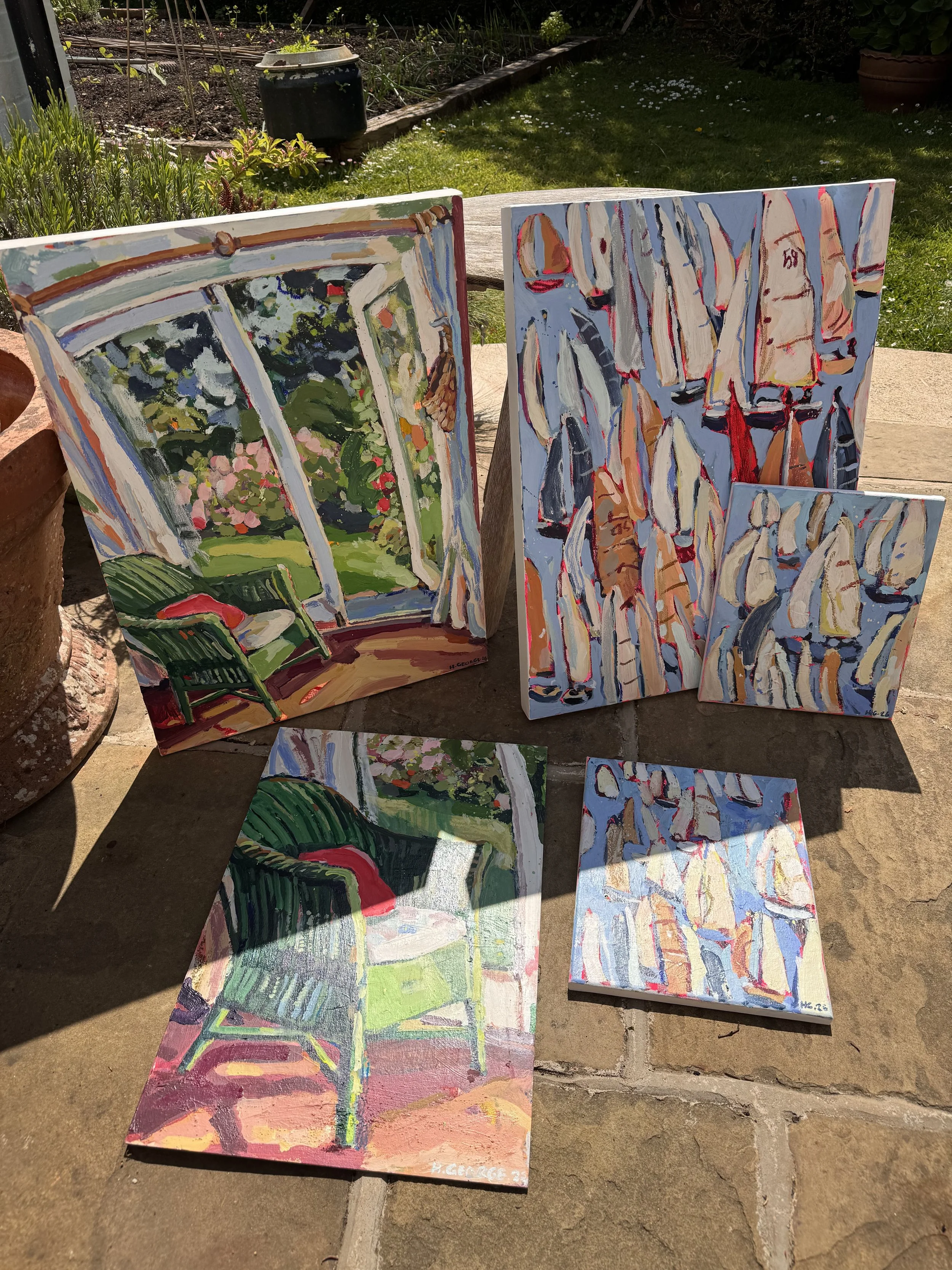 Several colorful paintings of still life scenes and landscapes displayed outdoors on the ground, with garden and patio elements in the background.