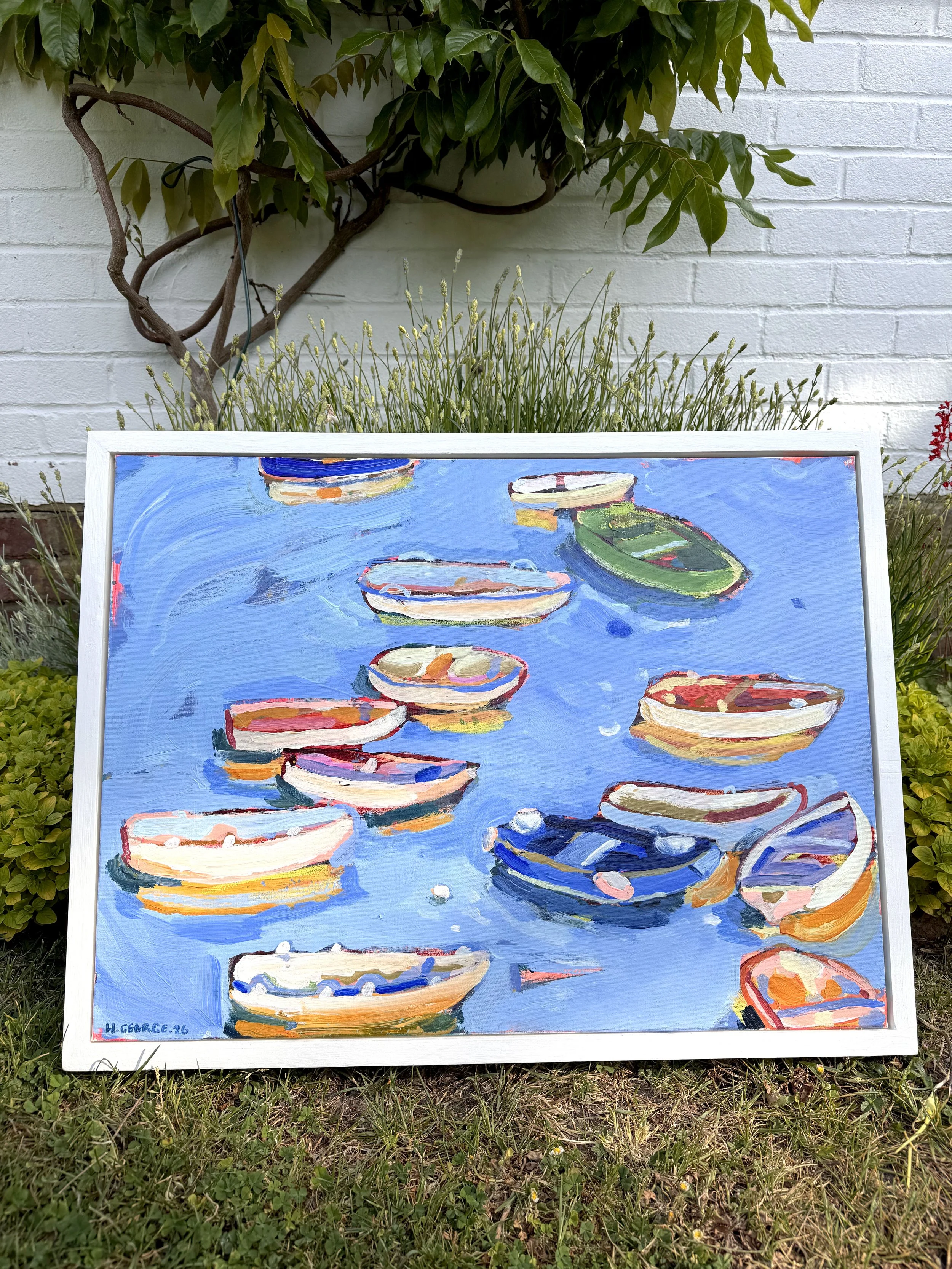 An outdoor scene with a white brick wall, green leafy vine, and purple flowers. A colorful painting of boats on water is propped against the grass in the foreground.