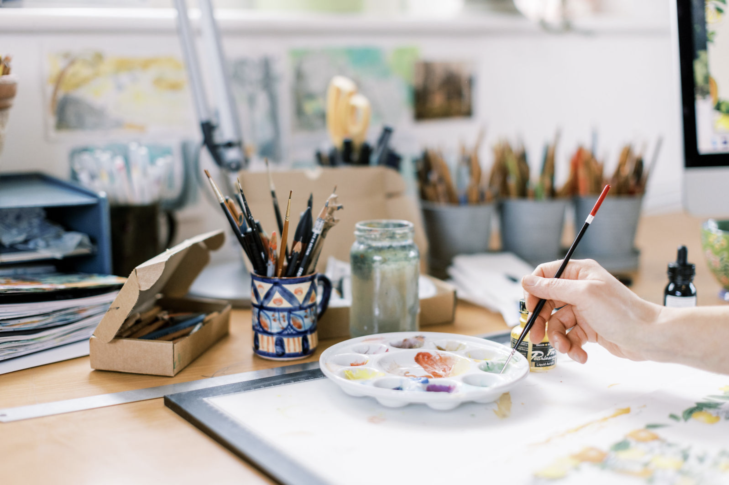 Hannah George - An artist's workspace with paintbrushes, a painter's palette with various colours, and art supplies on a wooden table.