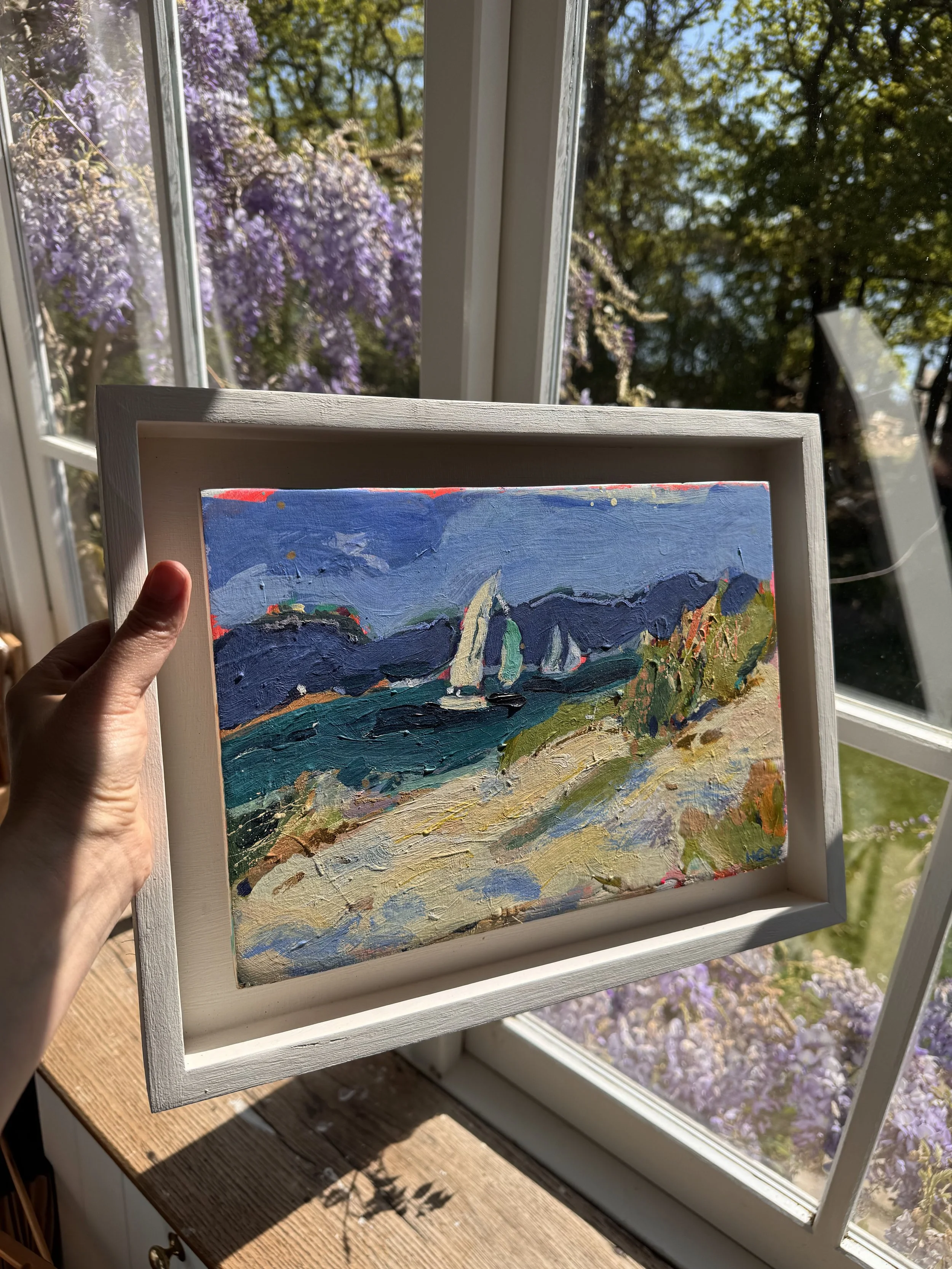 A person holding a small framed painting of a colorful landscape with mountains, trees, and a bright blue sky, in front of a window with purple flowers outside.