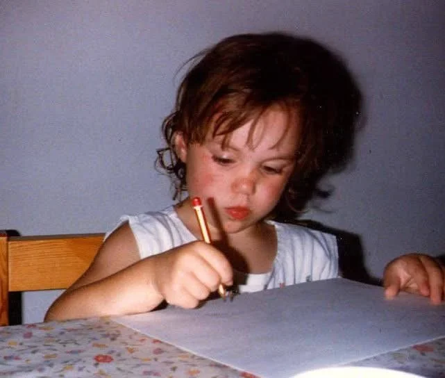 A young Hannah George sitting at a table, holding a crayon, appears to be drawing or colouring on a piece of paper.