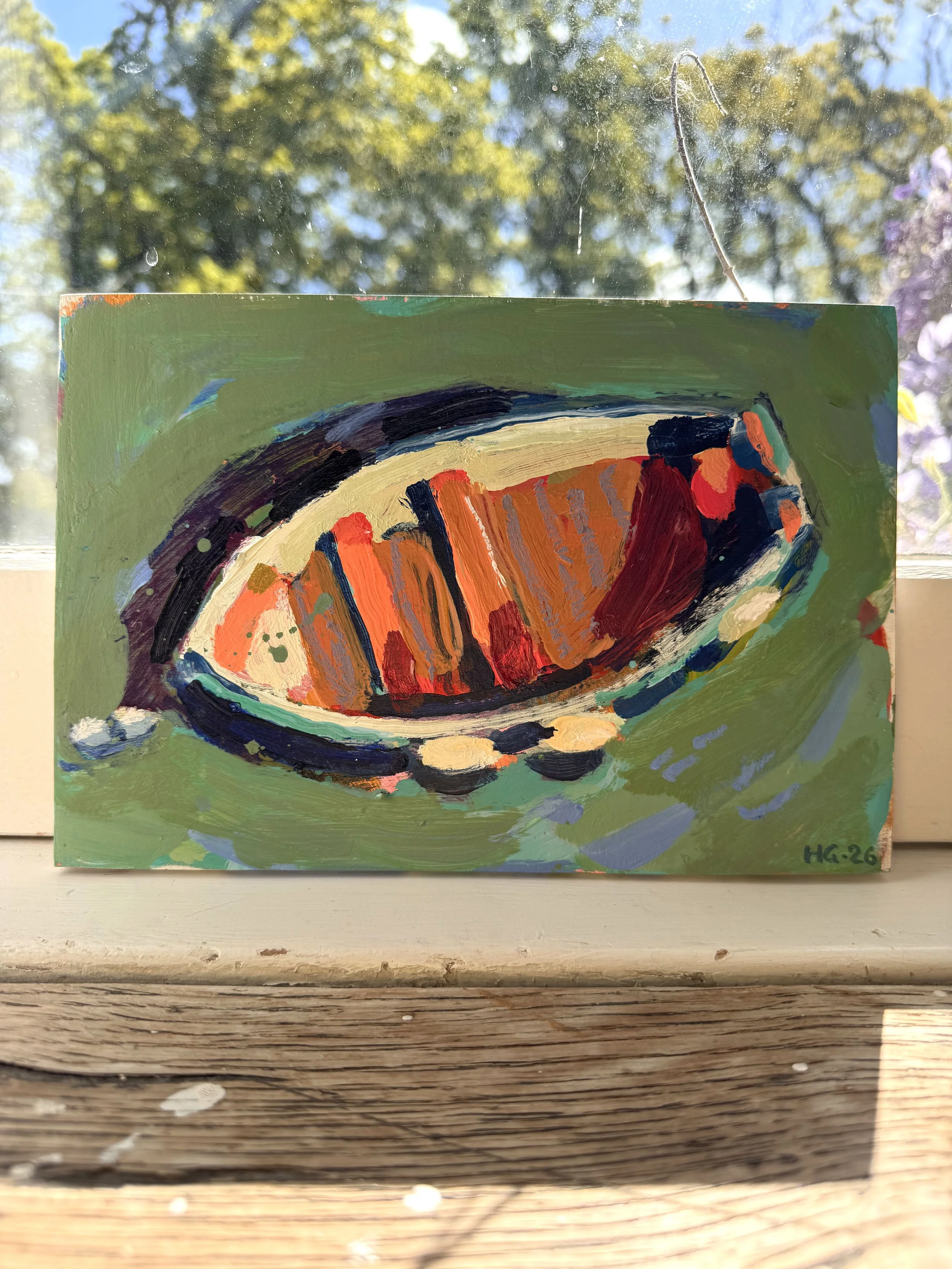 Close-up of an abstract painting on canvas featuring bold brushstrokes of orange, red, black, and blue, with a green background and the initials 'HG-26' in the bottom right corner. The painting leans against a window with a scenic outdoor view of tre