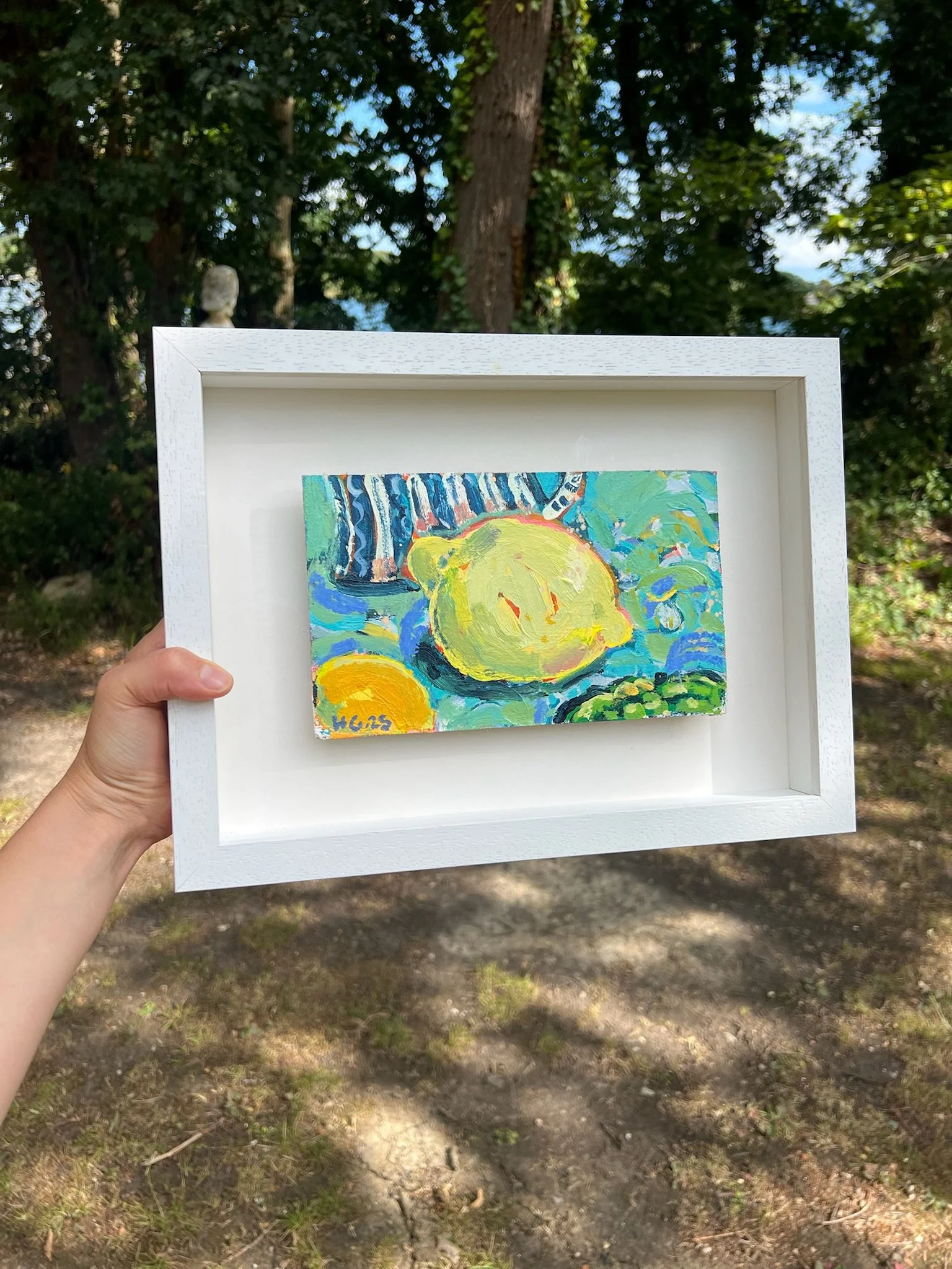 A person is holding a small framed painting outdoors in a wooded area, with trees and sky in the background. The painting features abstract, colorful shapes and textures.