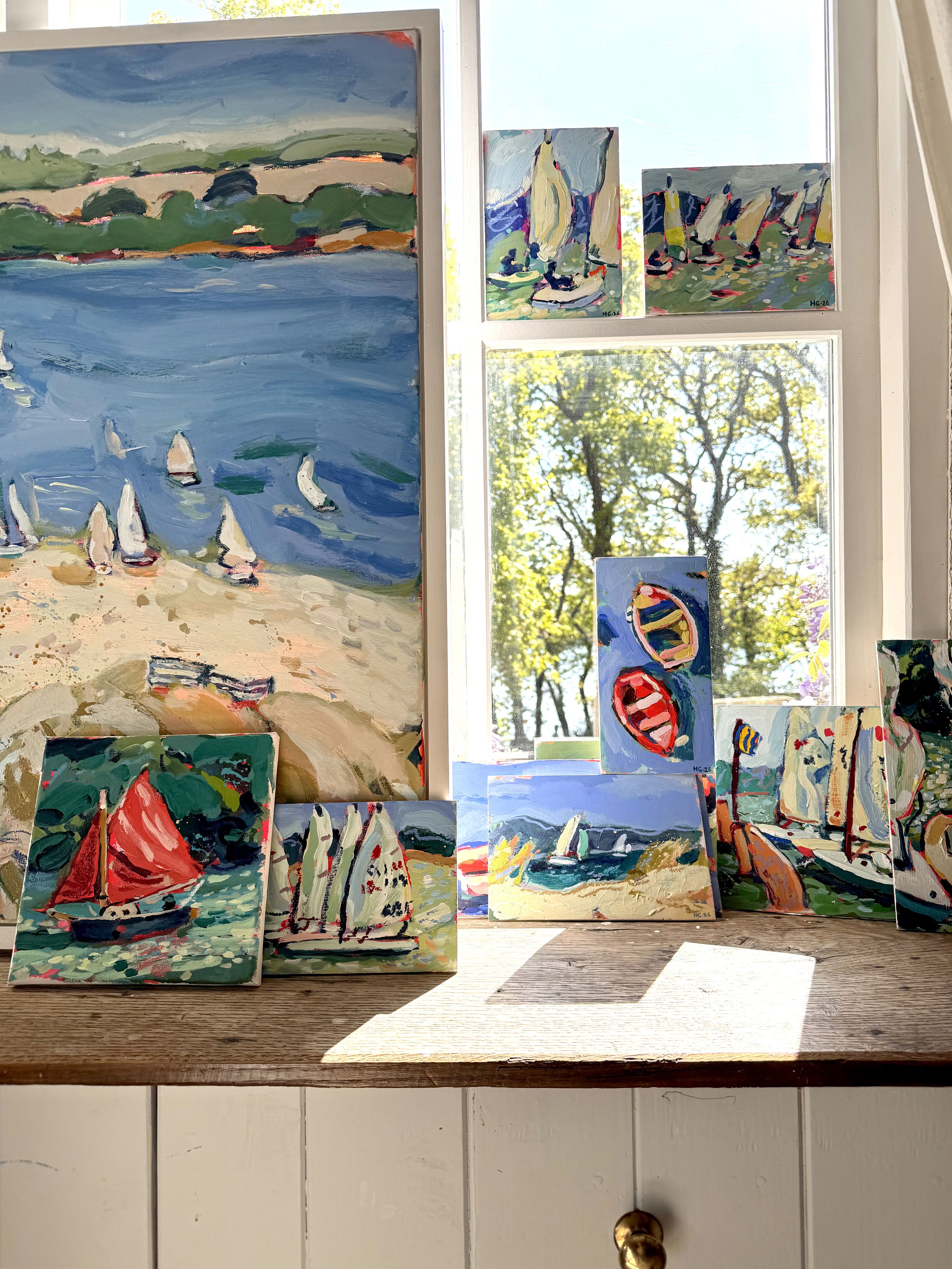 Set of colorful impressionistic paintings of boats and water scenes displayed on a shelf near a window with trees outside.