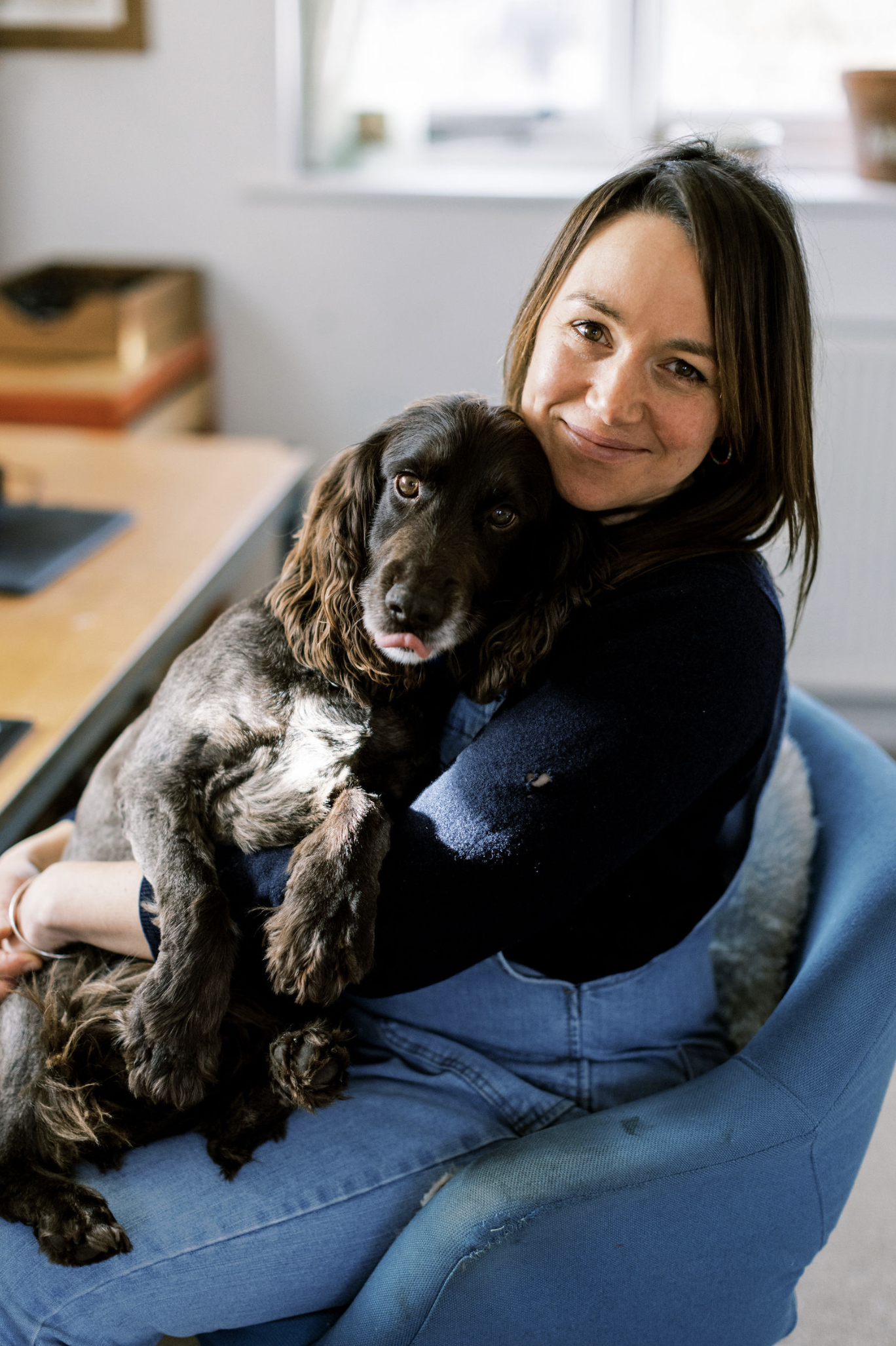 A woman sitting on a blue chair in a room, holding and hugging a long-haired brown and black puppy. The woman is smiling and looking at the camera, while the puppy is looking slightly off to the side.