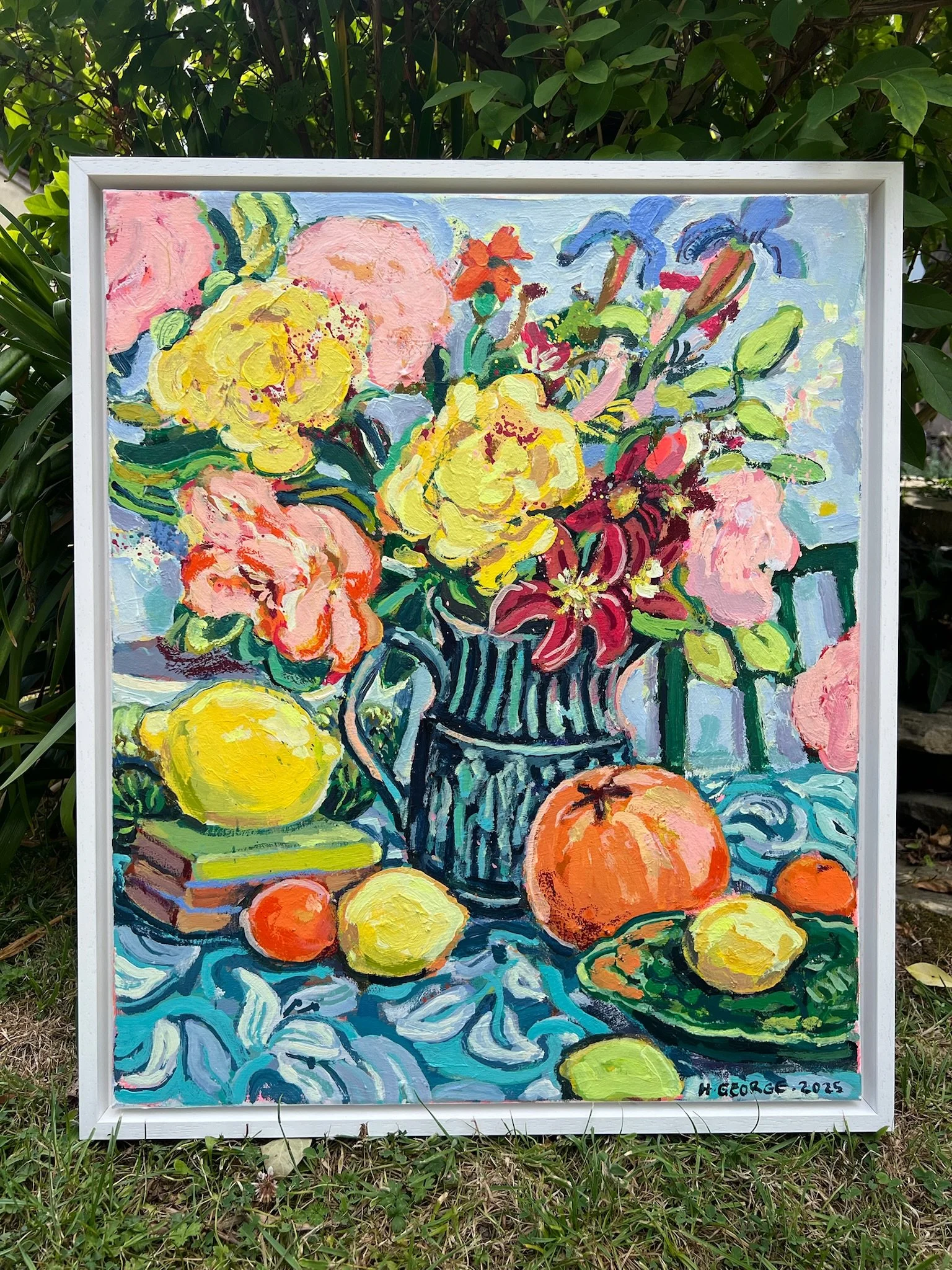 A colorful oil painting of a still life scene with a bouquet of yellow, pink, and red flowers in a striped vase, surrounded by lemons and oranges, and set against a blue background. The painting is signed "H. George 2025" in the bottom right corner.