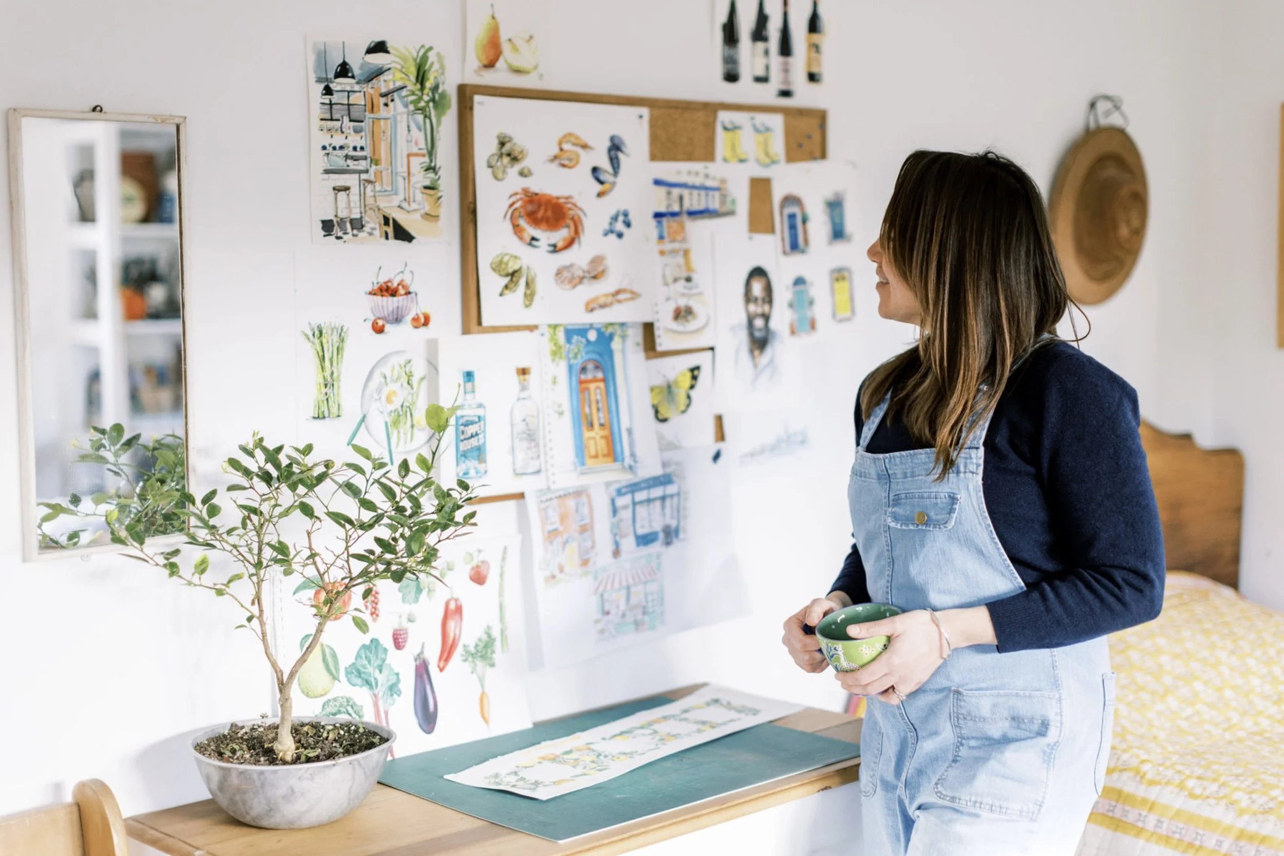 Hannah George, wearing a dark long-sleeve shirt and light blue denim overalls, standing in a bright room with watercolor illustrations of vegetables, animals, and objects on a wall. She is holding a green bowl and looking at the artwork.