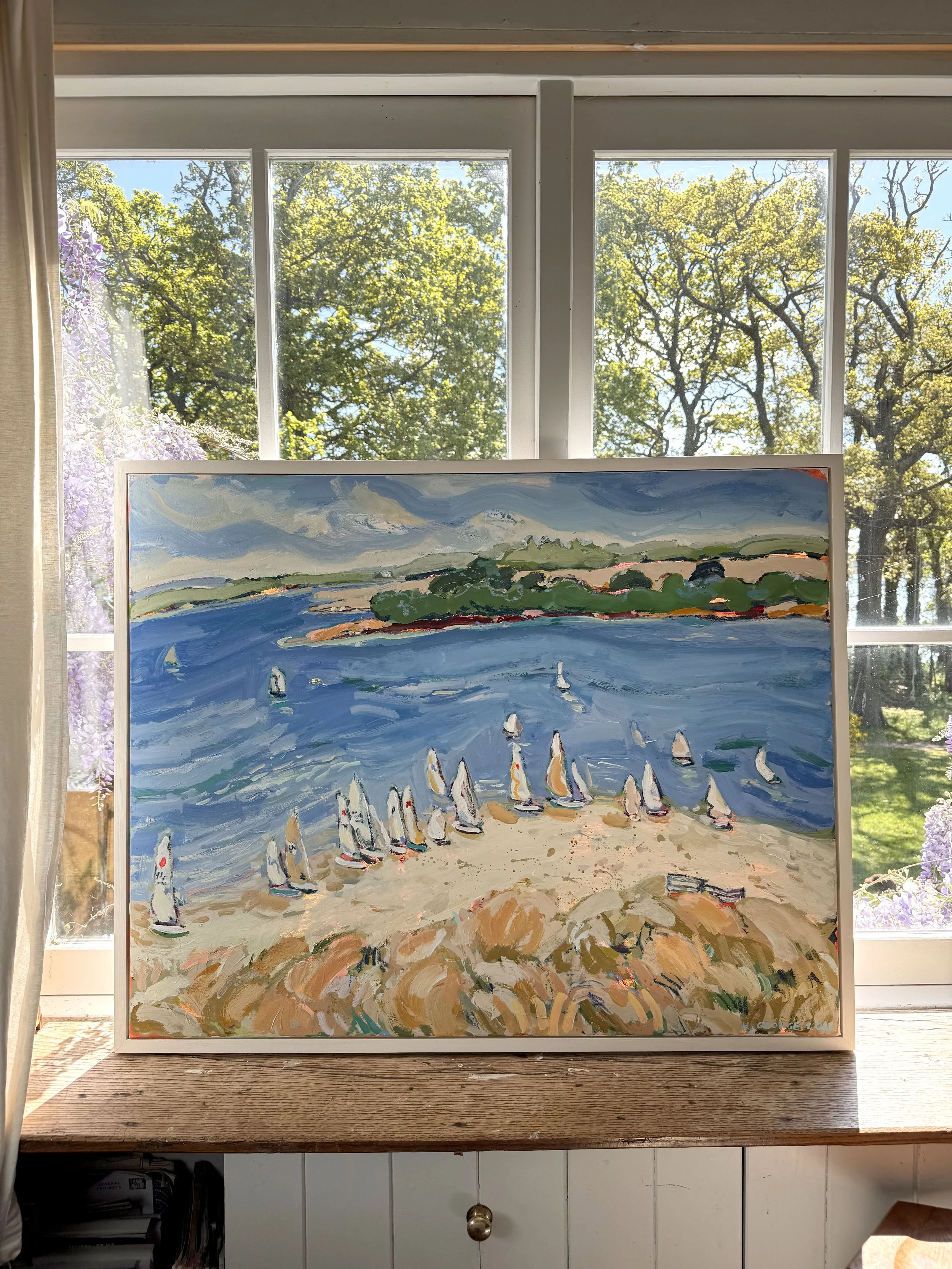 A painting of sailboats on the water along a shoreline with green hills and clouds in the sky, displayed against a window with green trees outside.