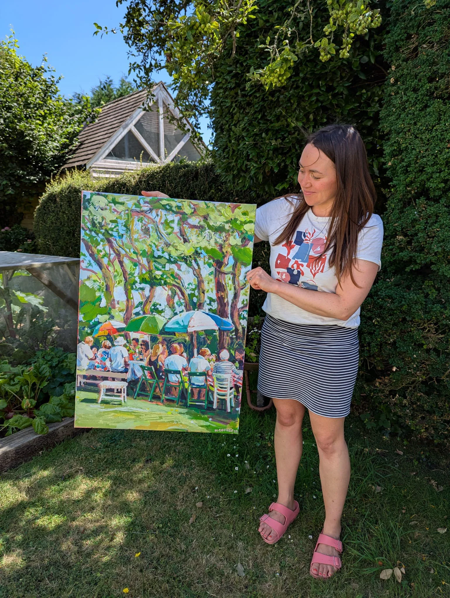 A woman with long dark hair wearing a white T-shirt with colorful graphic and a black-and-white striped skirt displaying a vibrant painting of an outdoor scene with trees and people on a grassy area, surrounded by green bushes and trees, with a small