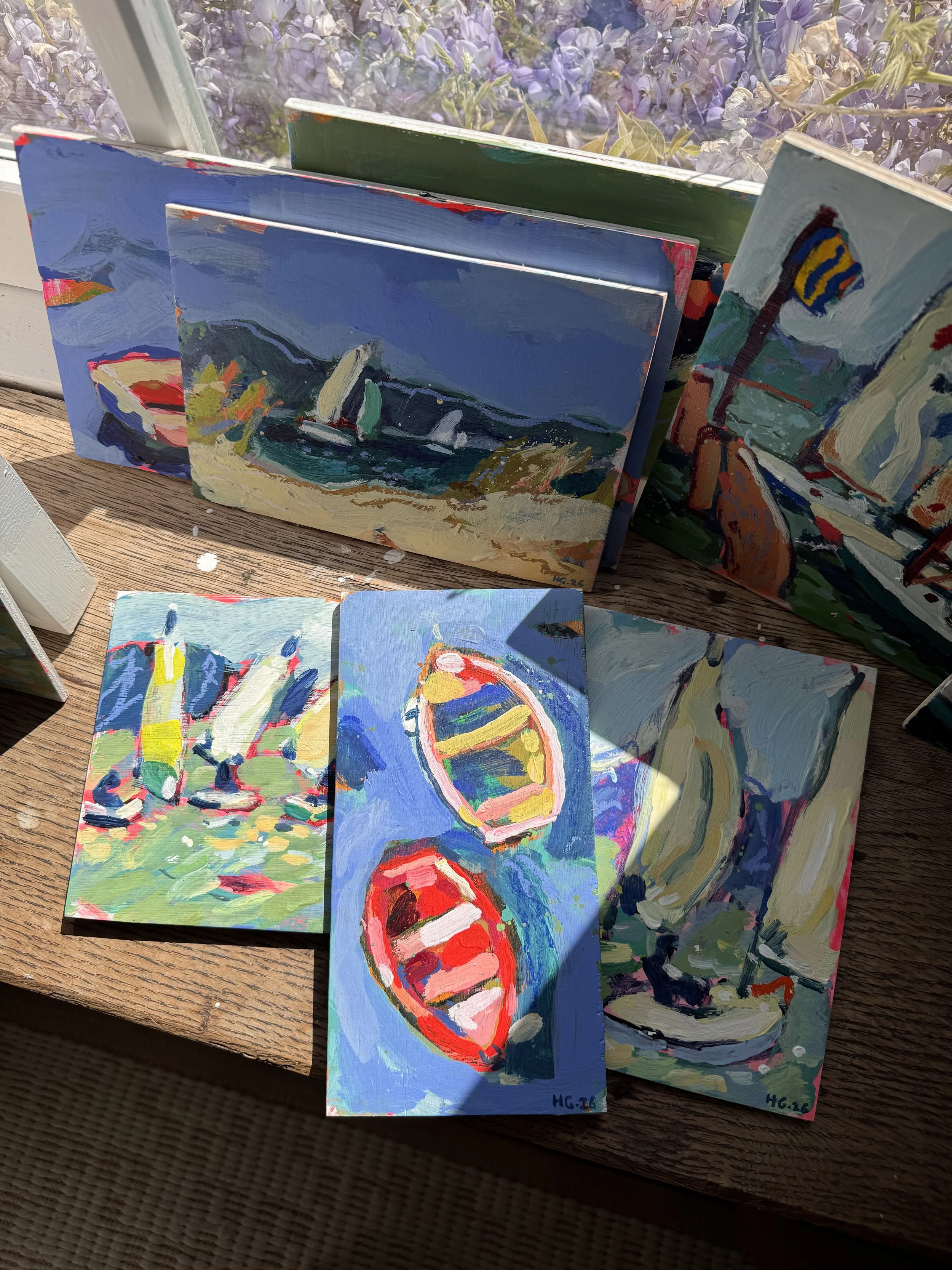 Multiple small, colorful, abstract paintings on paper, depicting various subjects including a sailboat, a face, and a flag, arranged on a wooden surface with a window and purple flowers in the background.