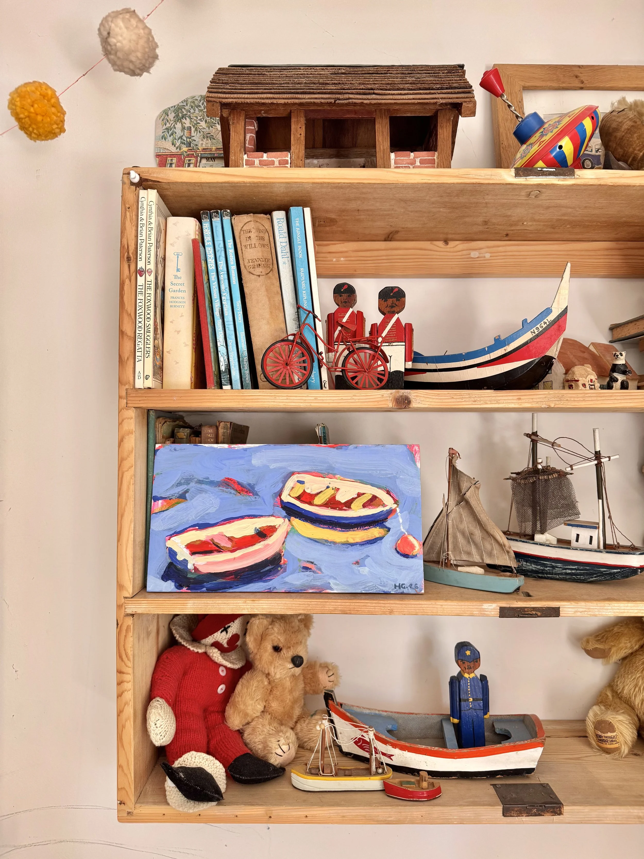 A wooden shelf with various decorative items including books, toys, and artwork. The top shelf has a small wooden house, a framed picture, and a colorful spinning top. The middle shelf has books, a wooden toy boat, and two carved wooden figures. The 