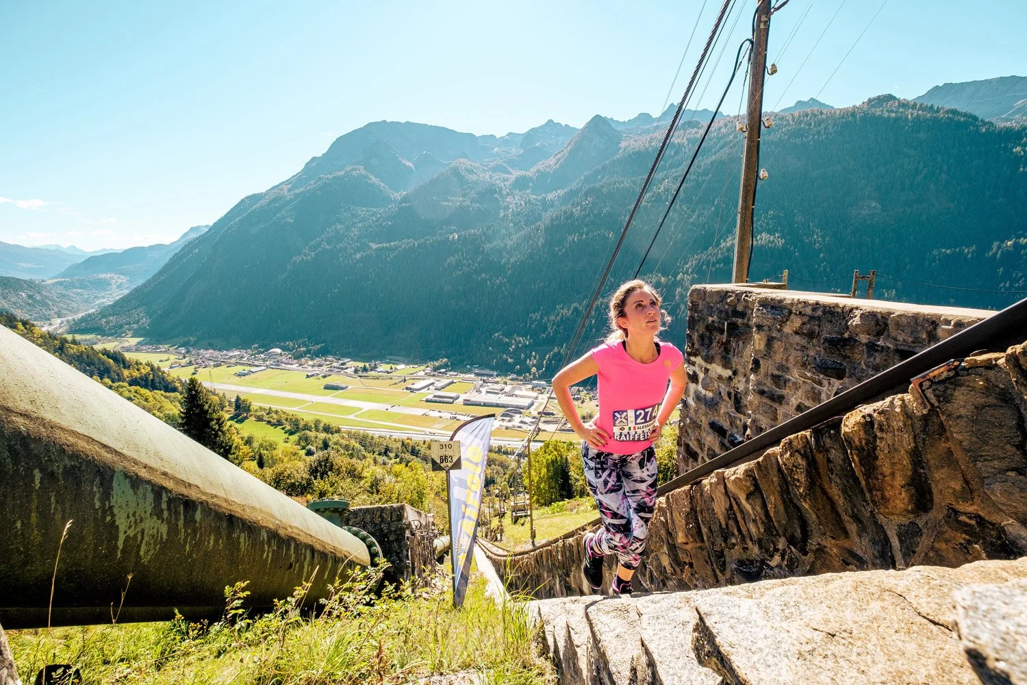 A woman running up stone stairs outdoors in a mountainous area during daylight, wearing pink shirt and patterned leggings, with a race bib and number 274.
