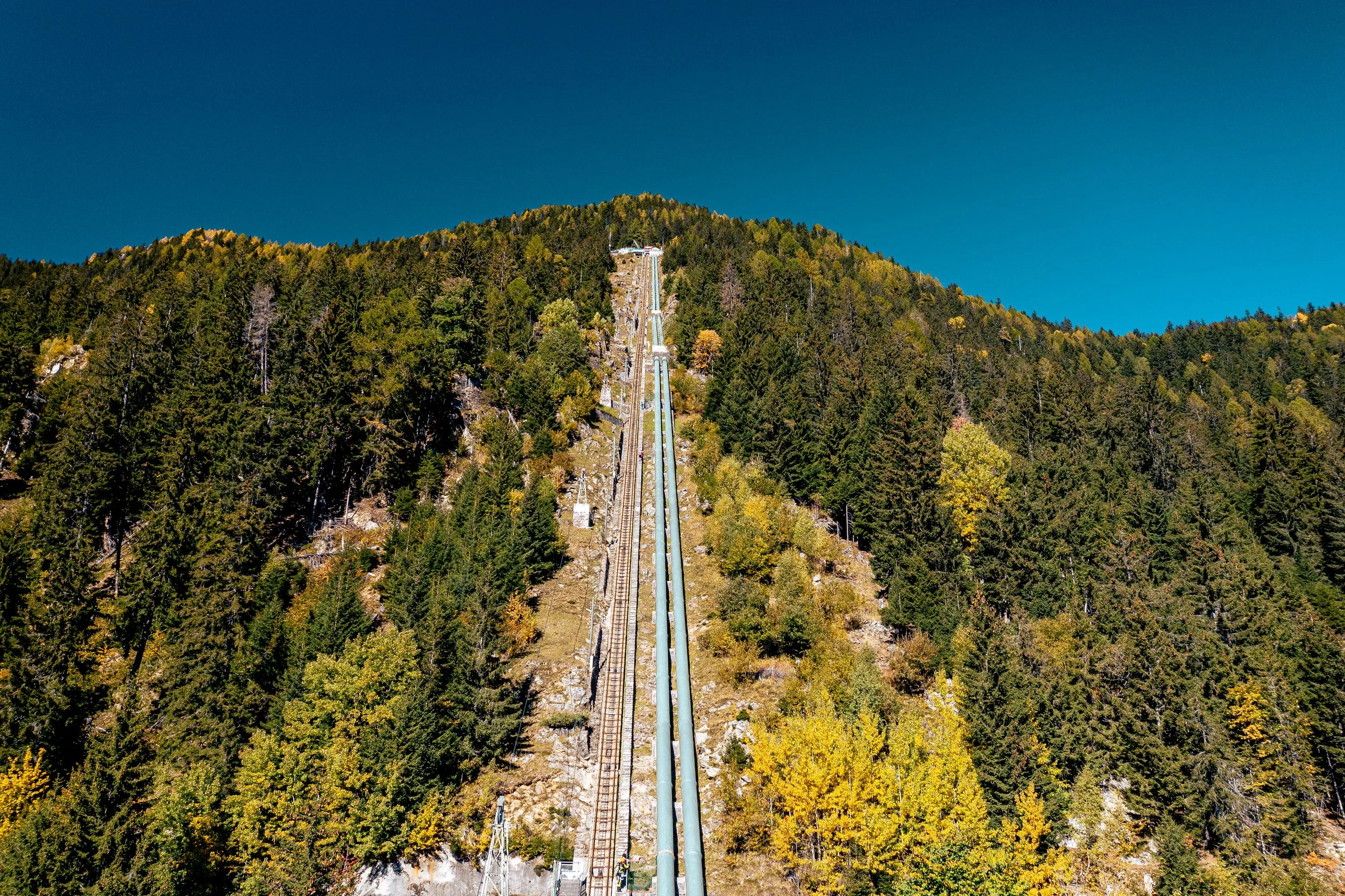 A hillside with a long ski jump ramp and a ski lift track, surrounded by trees with autumn foliage, against a clear blue sky.