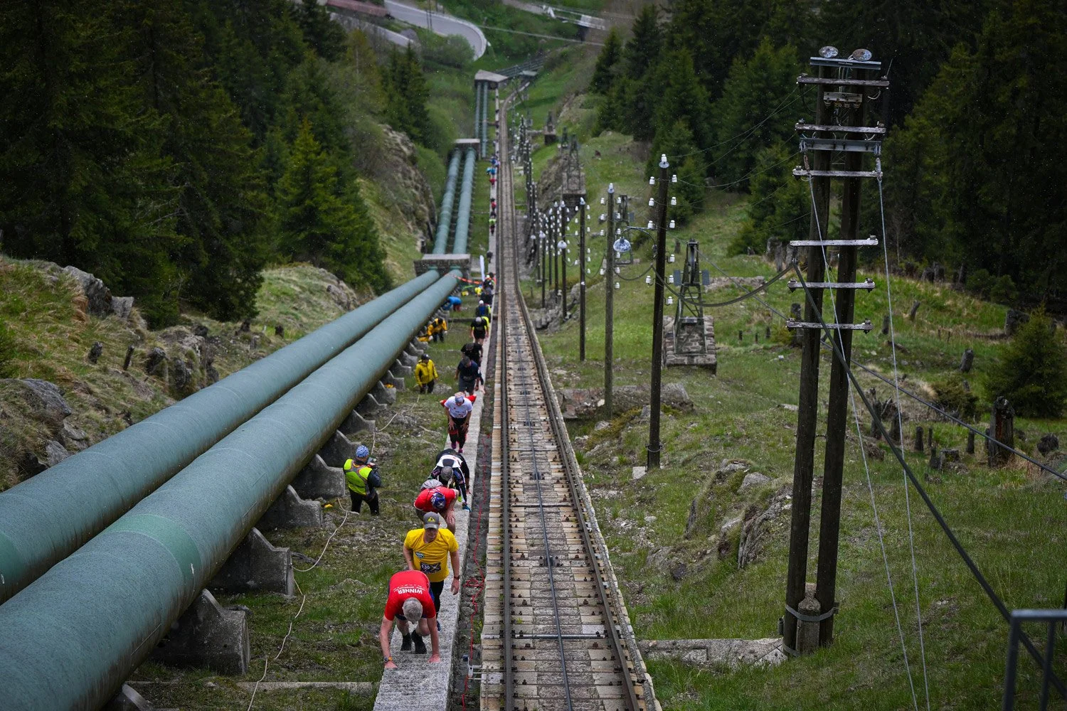 A group of hikers walking along a narrow trail next to large green pipes and electric poles in a mountainous area surrounded by trees.