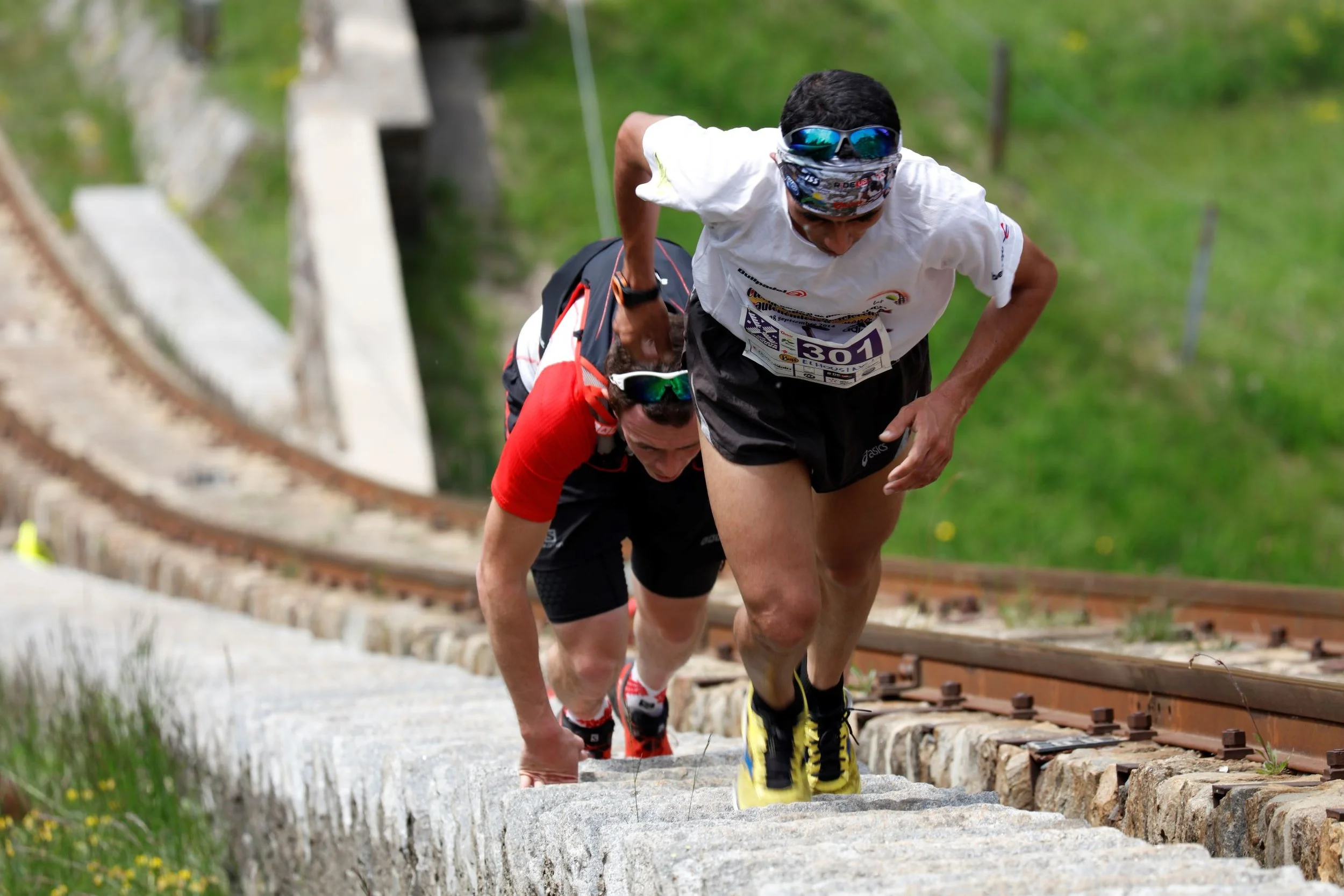Two male runners participating in a race, climbing a stone staircase next to a railway track with green grass and trees in the background.