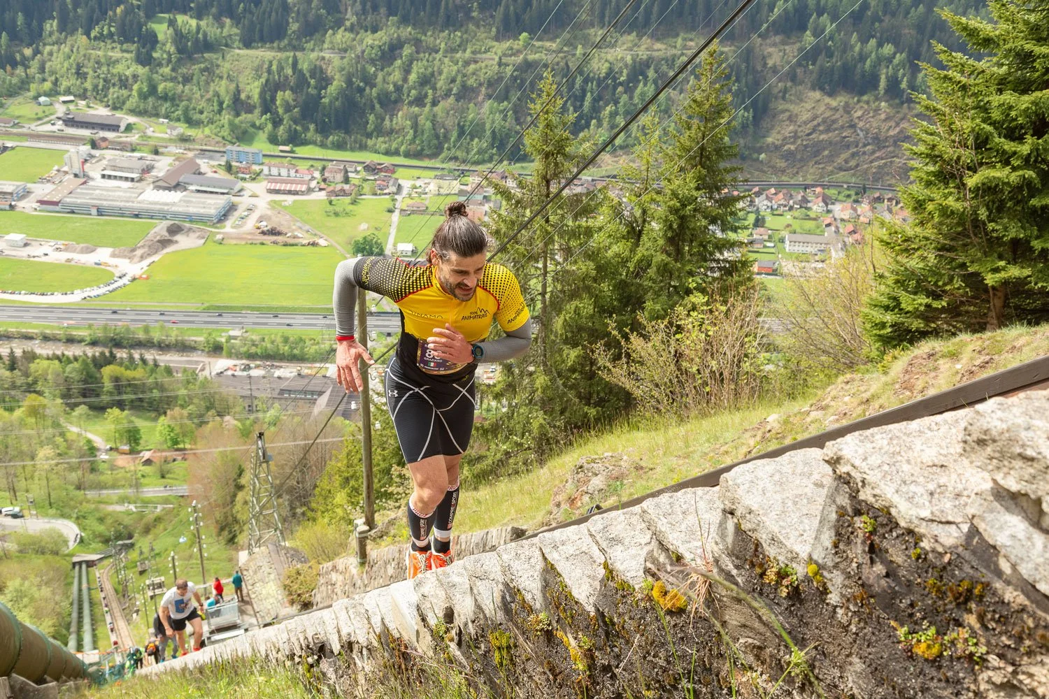 A male trail runner in a yellow and black athletic shirt and black shorts climbing a steep outdoor staircase made of stones, with a scenic view of a valley, trees, and hills in the background.