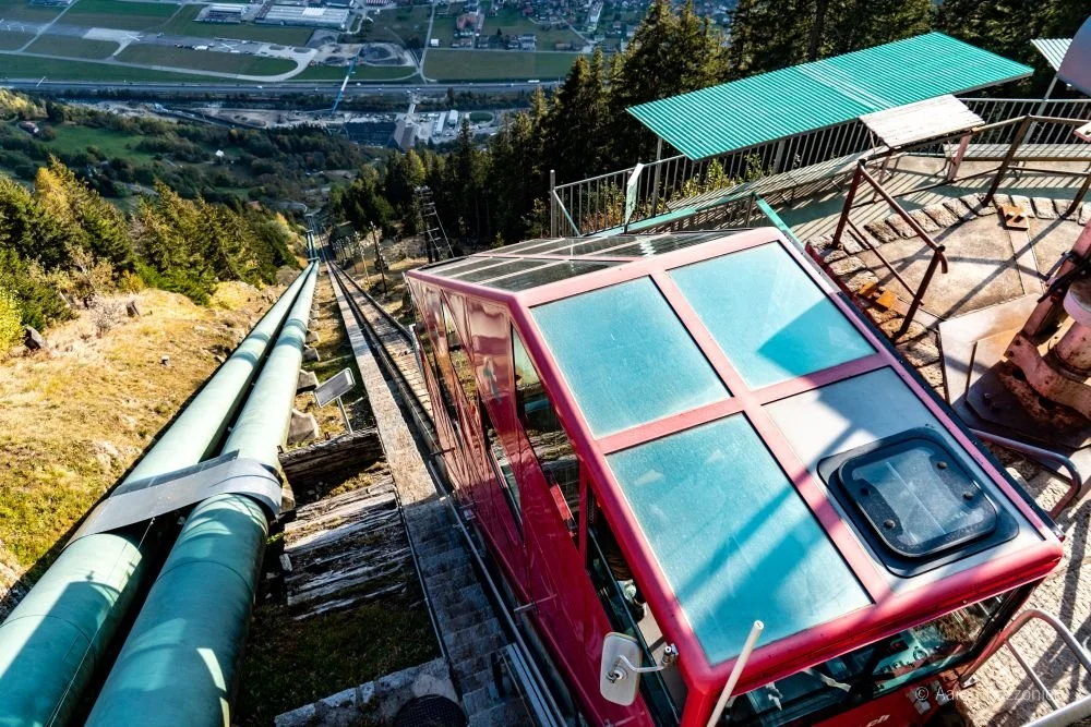 A ski lift cabin ascending a mountain with green pipelines running alongside it, overlooking a valley with buildings and roads.
