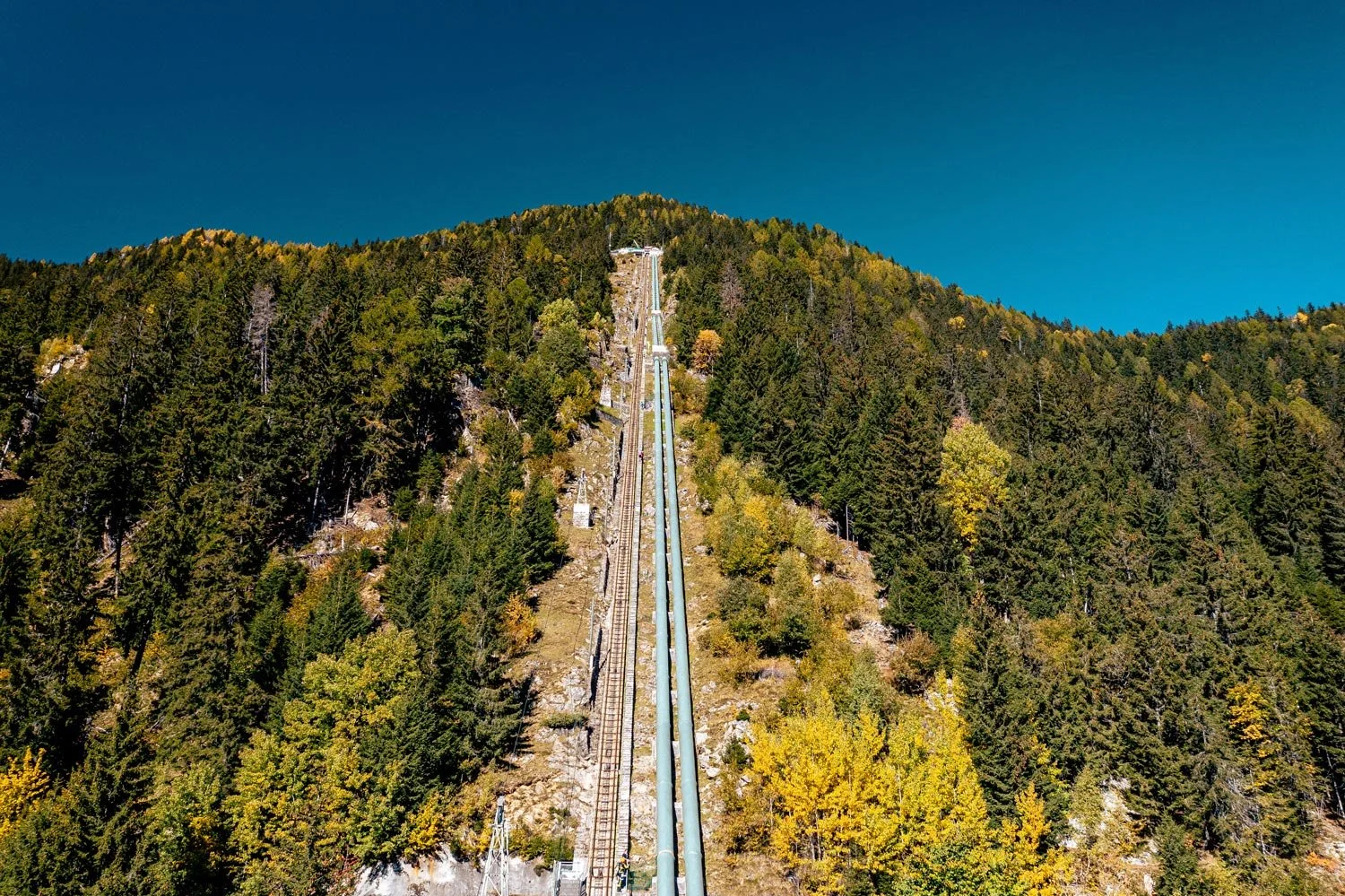 Ski jump ramp on a steep hillside covered with green and yellow trees, leading up to a building at the top, under a clear blue sky.