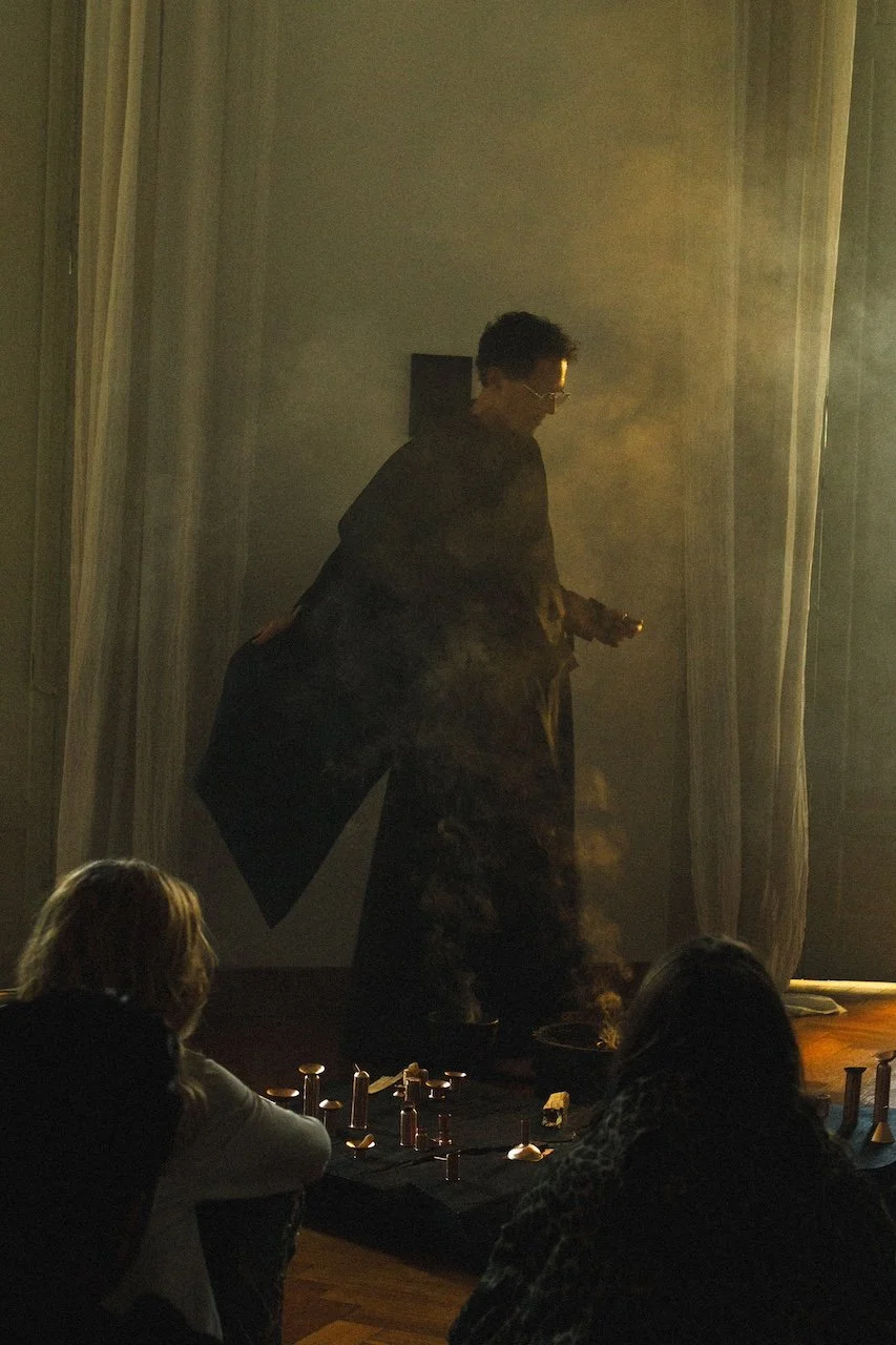 A person on stage, possibly a magician or performer, is surrounded by smoke and standing in front of an audience in a dimly lit room. The audience members are seated at a table with small objects and are watching the performance.