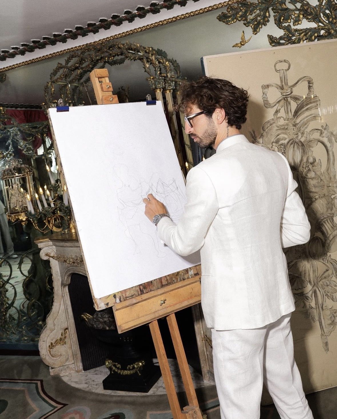 A man dressed in a white suit with glasses is drawing on a large white canvas positioned on an easel. The setting appears to be an ornate, elegant room with gold decorative elements, a fireplace, candelabras, and a painting of a sword with an ornate handle in the background.