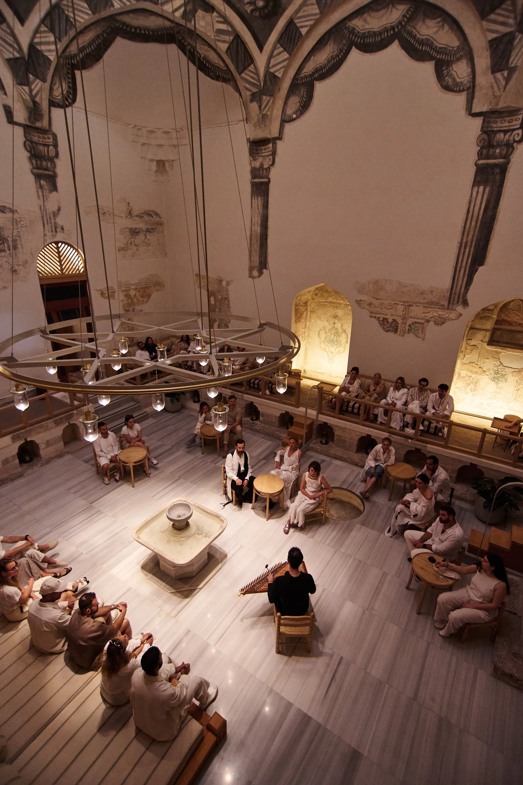 A group of people in white attire sitting around a central musician playing a traditional stringed instrument, in a spacious room with a high ceiling, frescoed walls, and a modern circular chandelier.