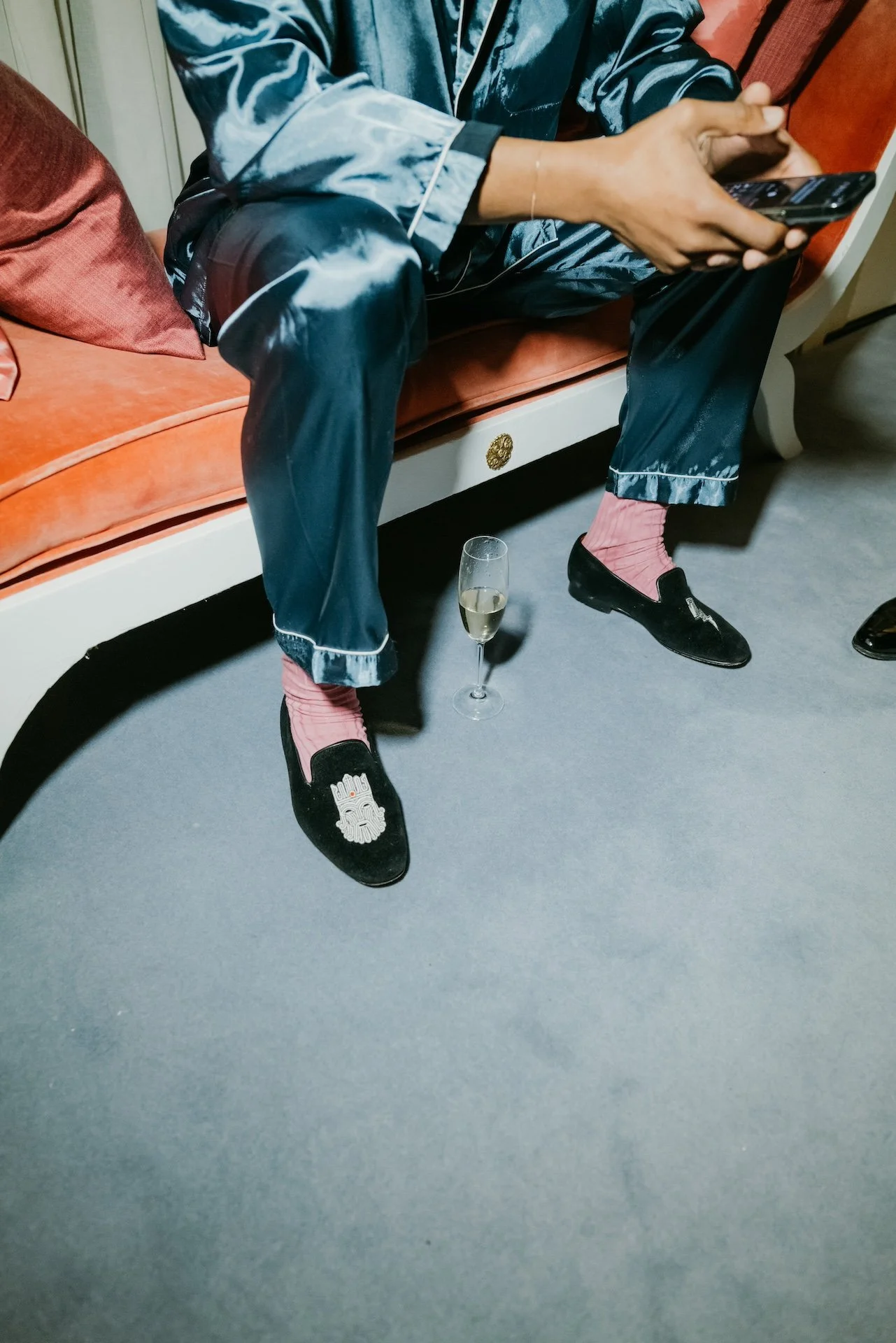 A person sitting on a vintage sofa, wearing shiny blue pajamas, pink socks, and black slippers with a white tiger face. They are holding a phone, and a glass of champagne is on the floor nearby.