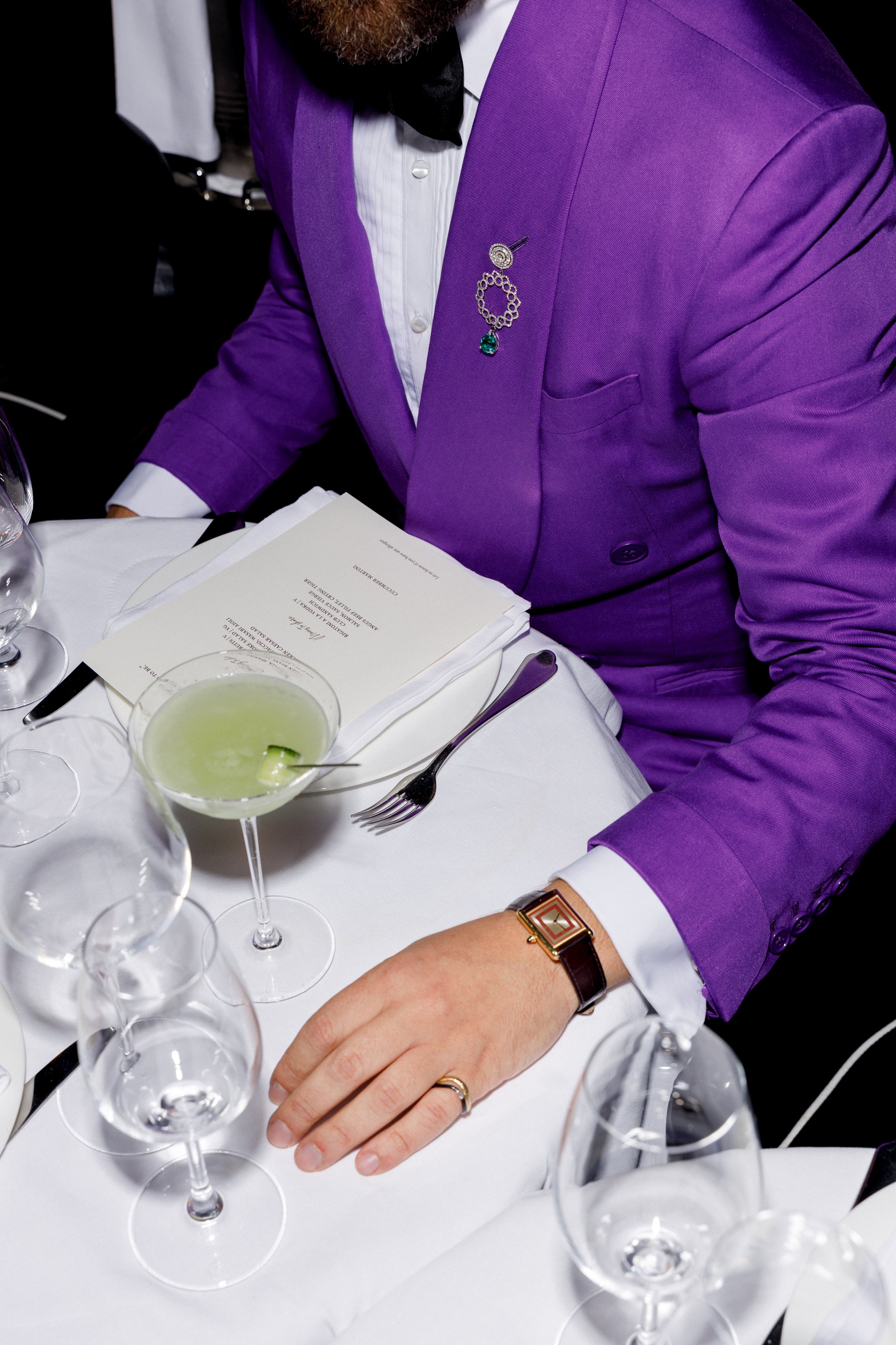 A man dressed in a purple tuxedo with a white shirt, black bow tie, and jewelry, sitting at a dining table with a menu and various glasses of drinks, including a green cocktail garnished with a cucumber slice.