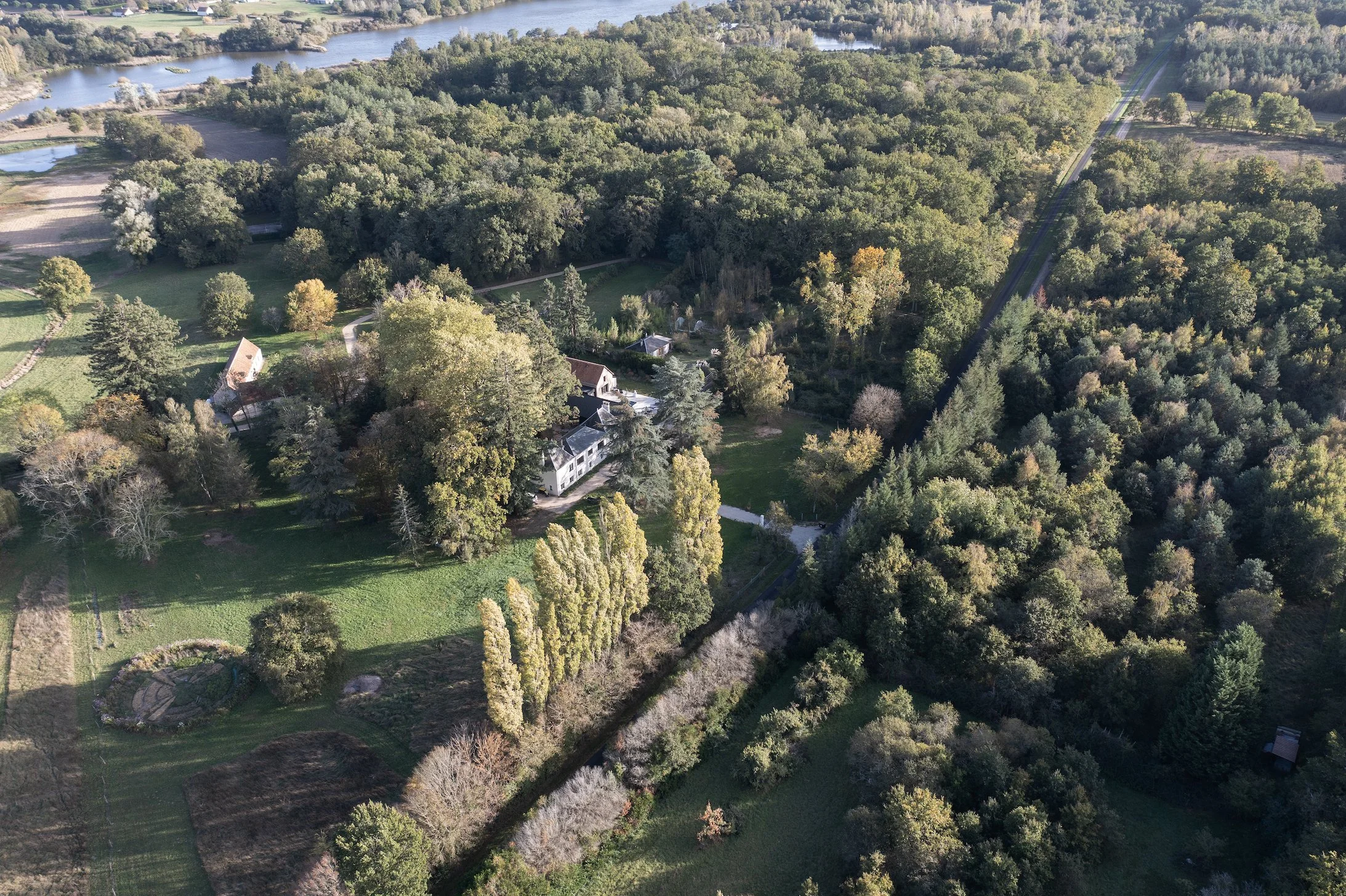 Aerial view of a house surrounded by trees and greenery, with a river nearby and a road running through the area.