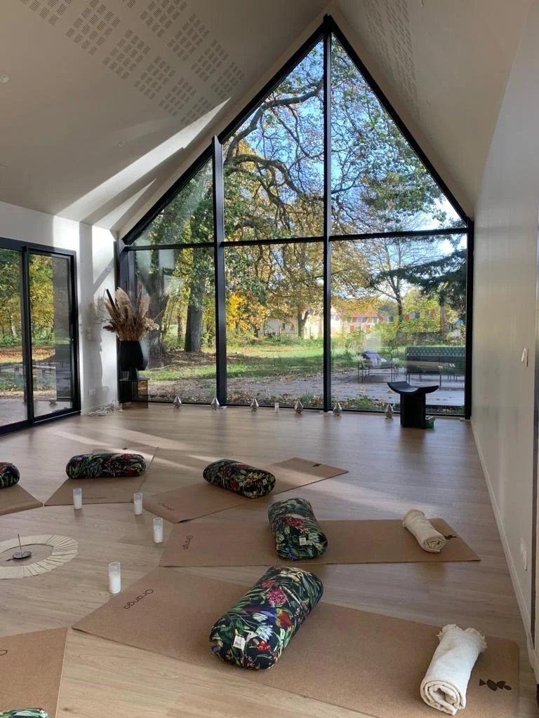 A serene yoga or meditation space with large glass triangular windows, wooden floors, and yoga mats with bolsters and candles. Outside scenery includes trees and a park.