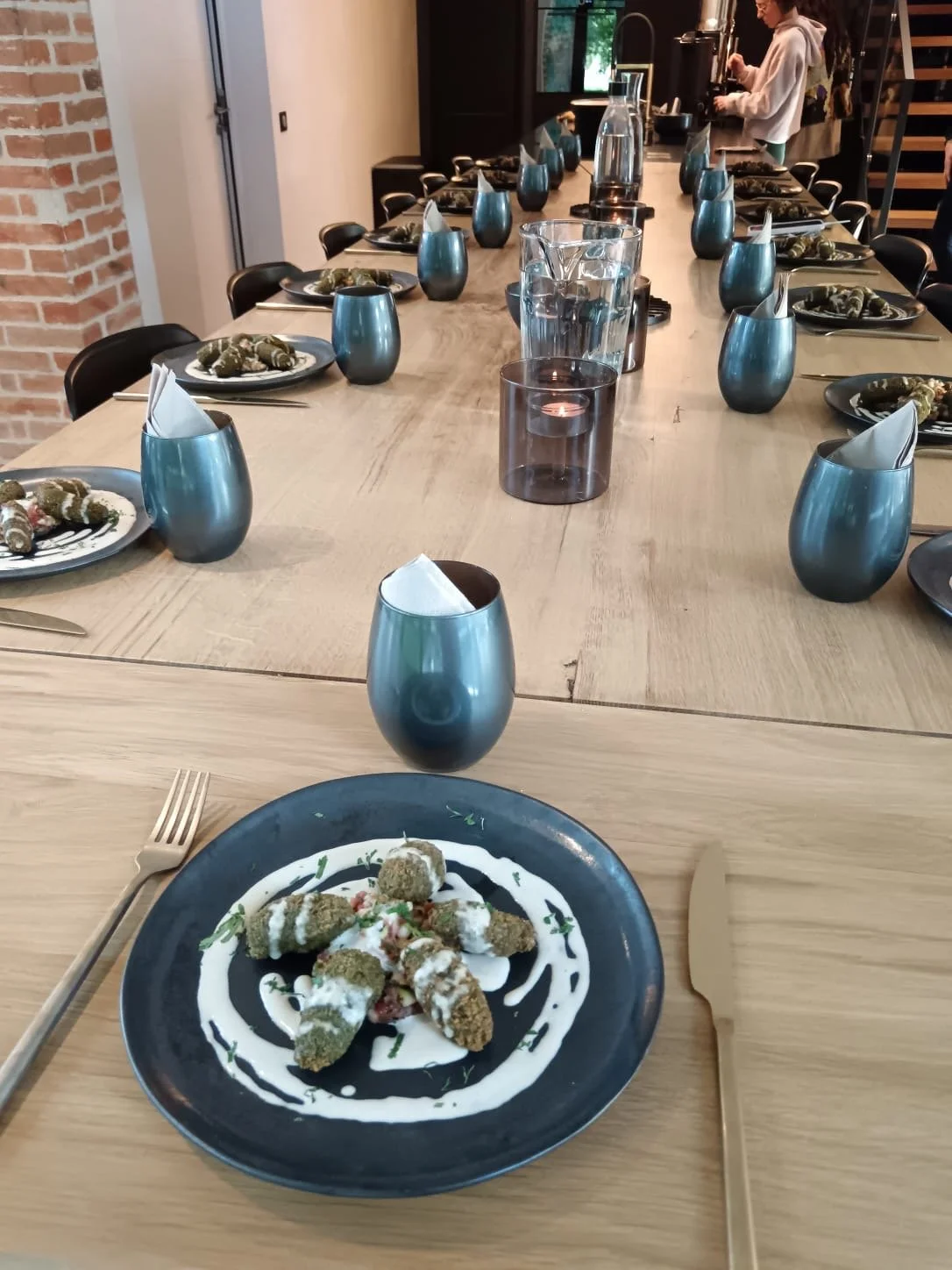 A dinner table set with plates of food, teal-colored cups, water glasses, and candles. There are plates with a dish that looks like stuffed or rolled items drizzled with sauce. The table is in a modern dining space with brick and white walls, and peo