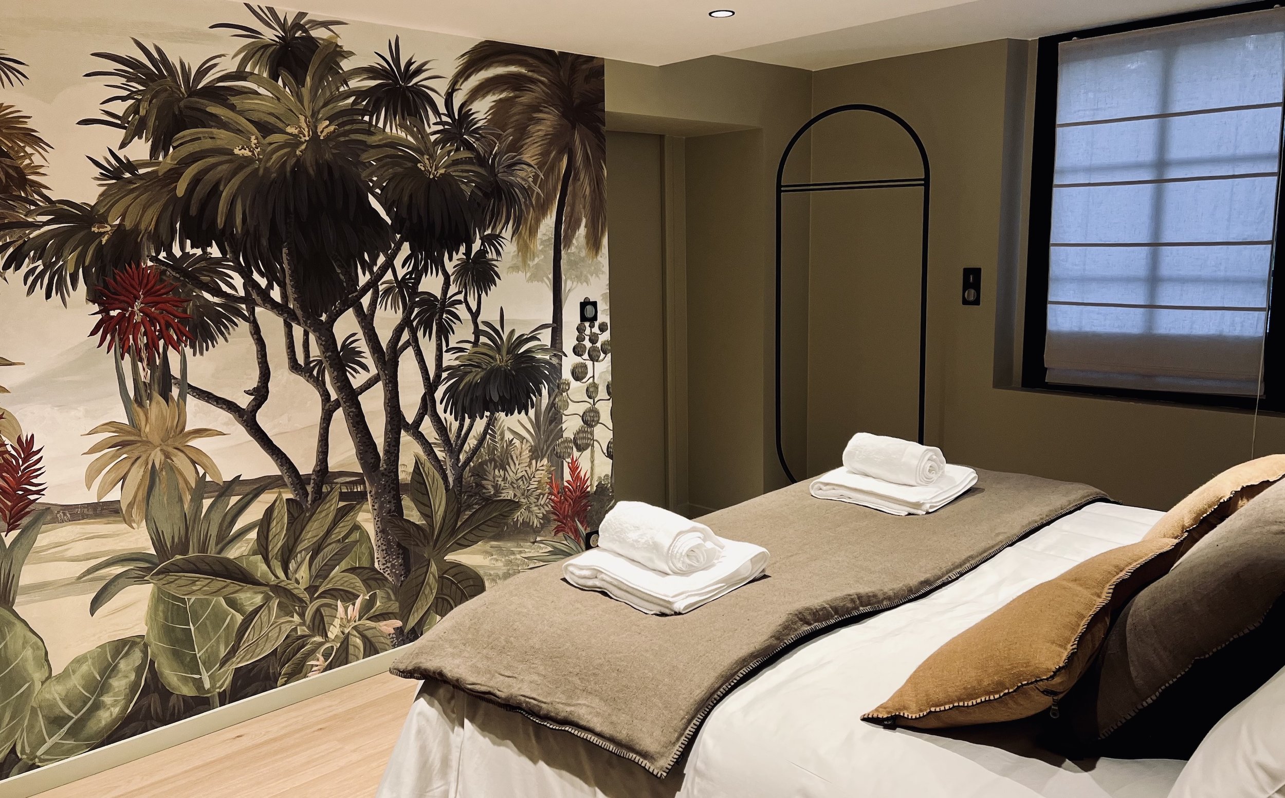 A bedroom with a jungle-themed mural of trees and foliage on the wall, a bed with neatly folded towels and pillows, a window with a white shade, and a black standing frame.