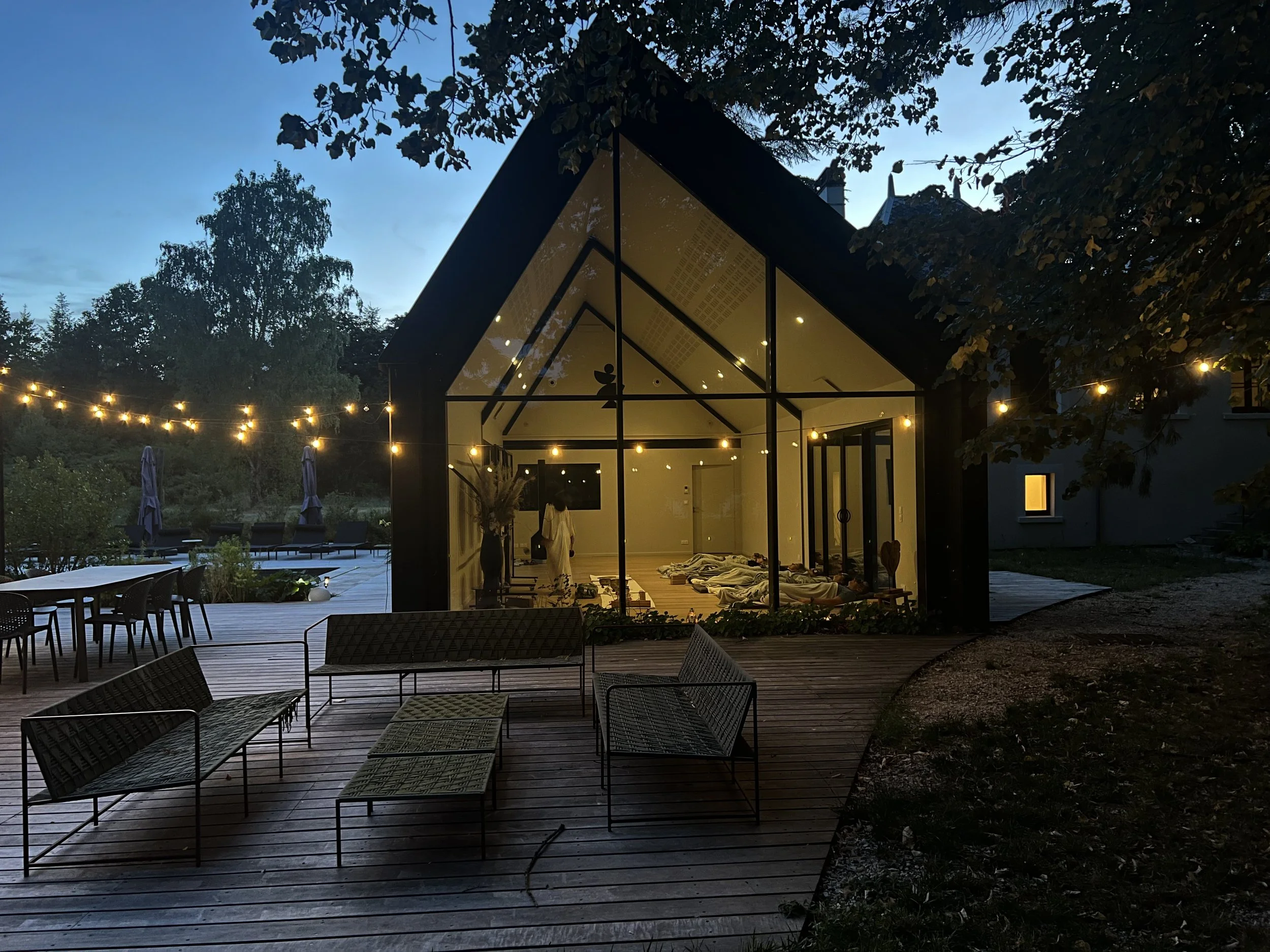 A glass-walled house with a pitched roof, illuminated from inside, at dusk. The house is on a wooden deck with outdoor furniture, string lights, and trees surrounds the area.