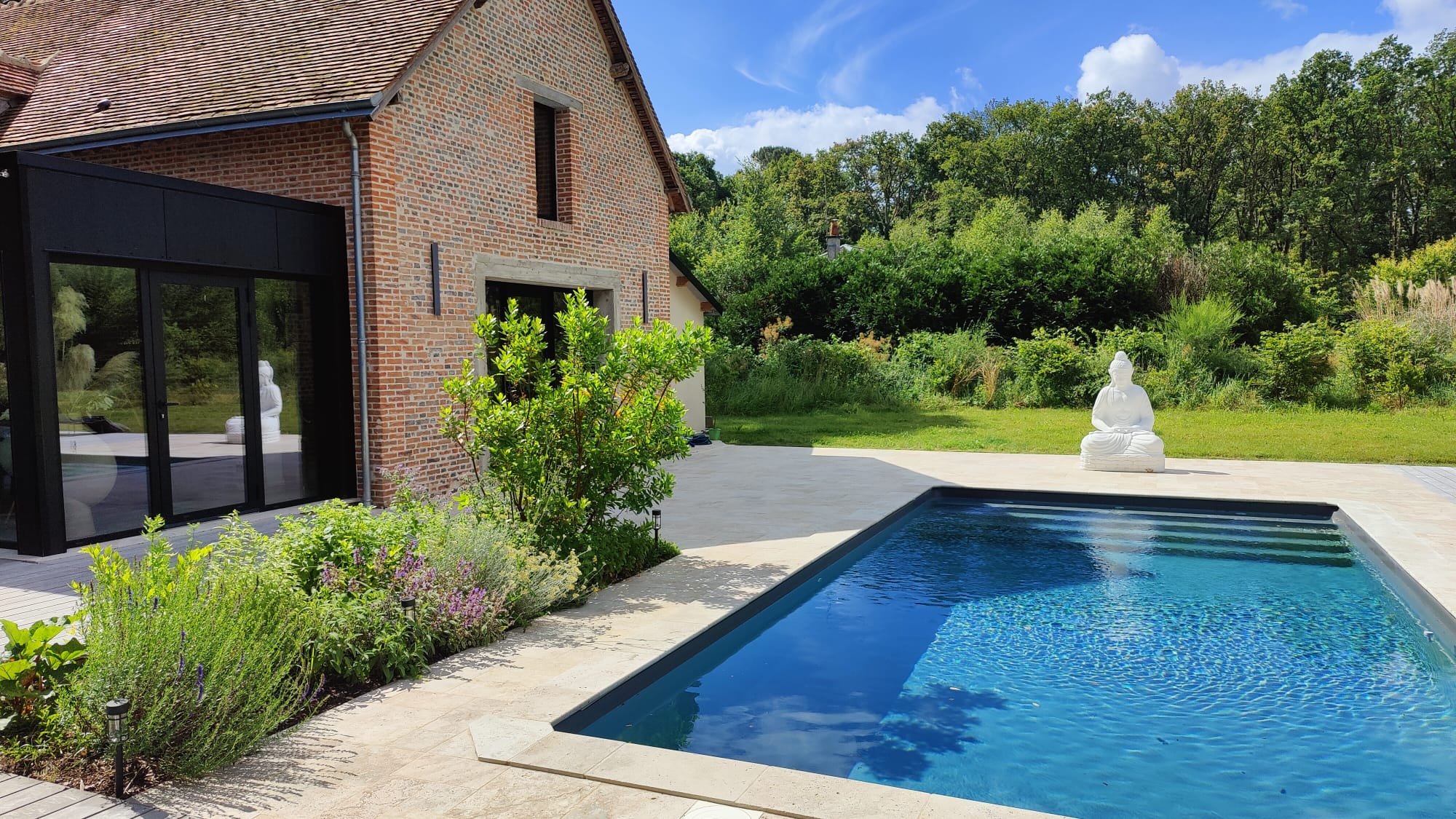 A backyard with a rectangular swimming pool, a Buddha statue on the patio, a garden with green bushes and flowers, a brick house with large black-framed windows, and a wooded hillside in the background under a partly cloudy sky.