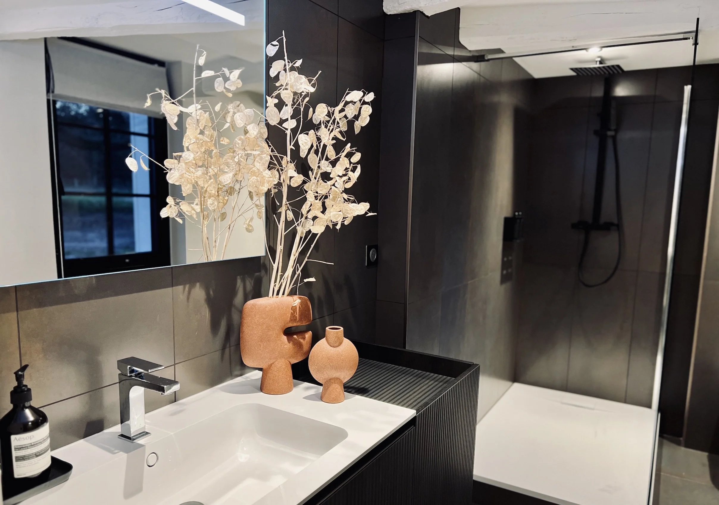 Modern bathroom with a black vanity, white sink, black faucet, decorative vases, and a large mirror. In the background, there is a walk-in shower with black walls and a showerhead.